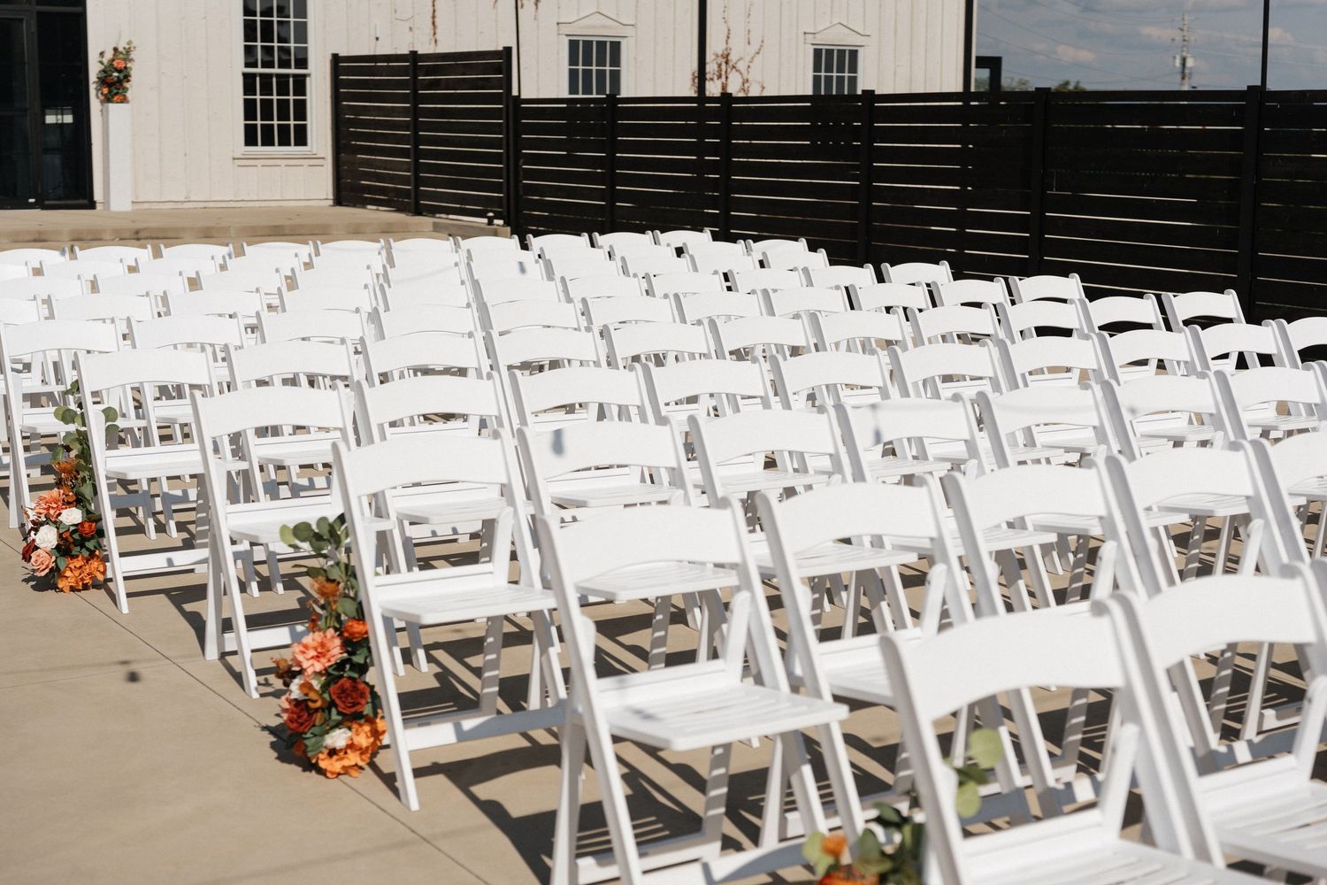 Rows of white folding chairs set up for outdoor wedding ceremony with orange floral aisle decorations.