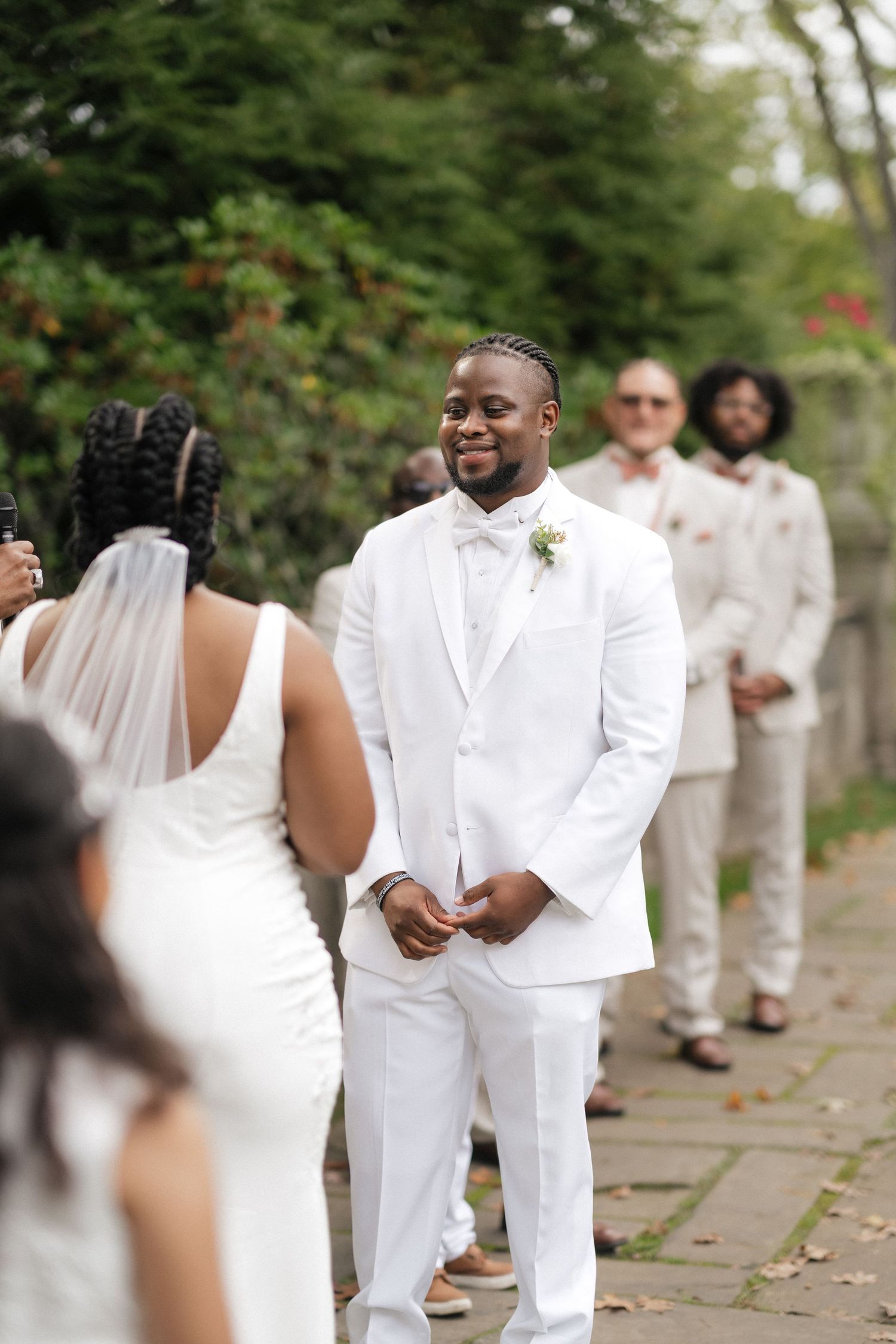 A wedding party stands outdoors in coordinated white attire against a backdrop of greenery and trees.