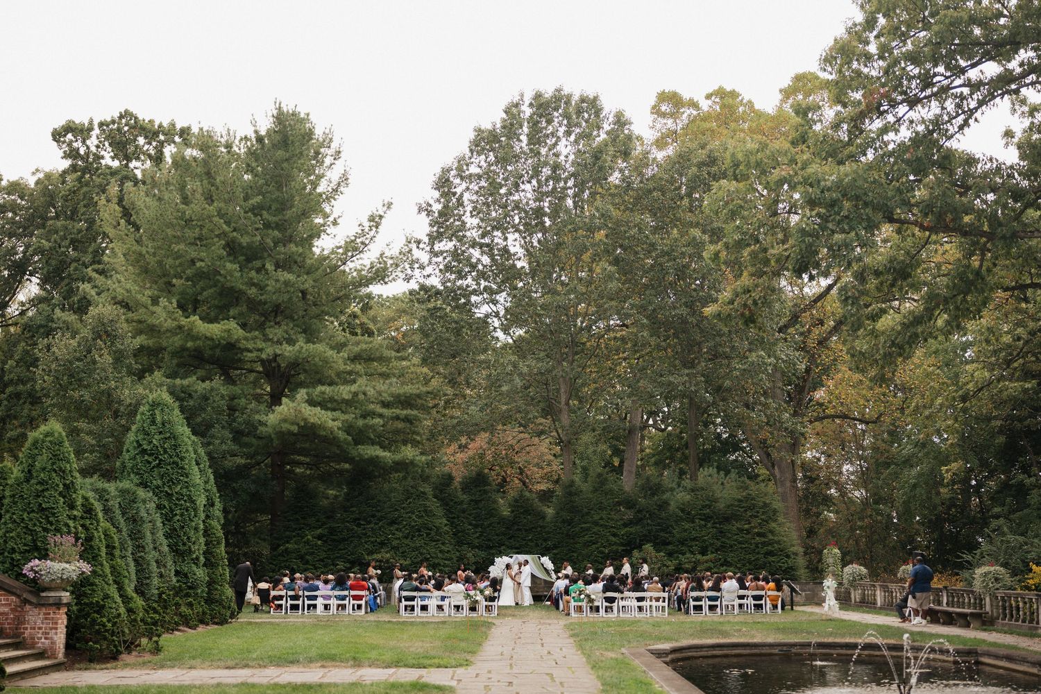 Wide panoramic view of an outdoor wedding venue with guests seated on white chairs.