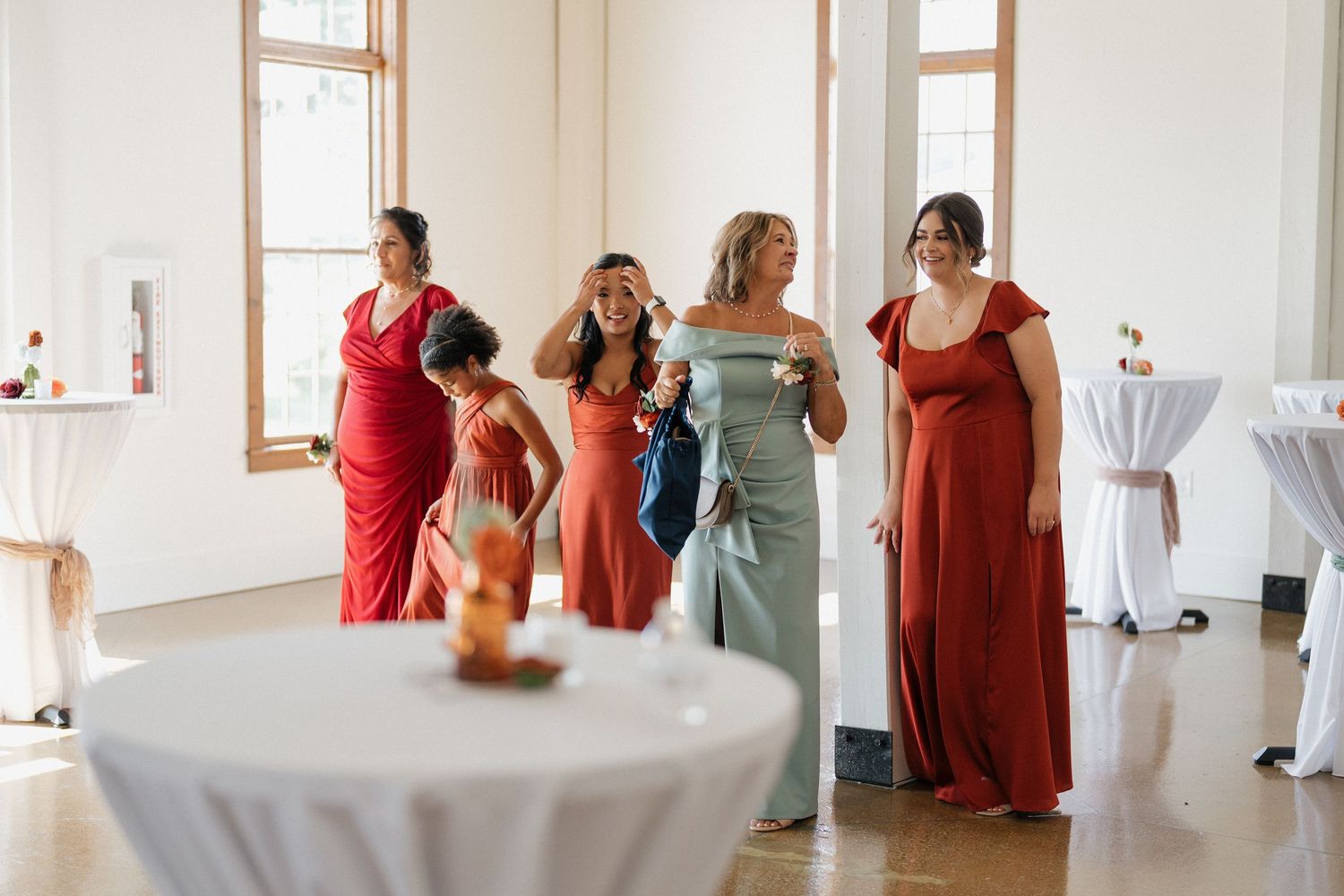 Wedding party members in burgundy and sage dresses gather in a bright indoor venue.