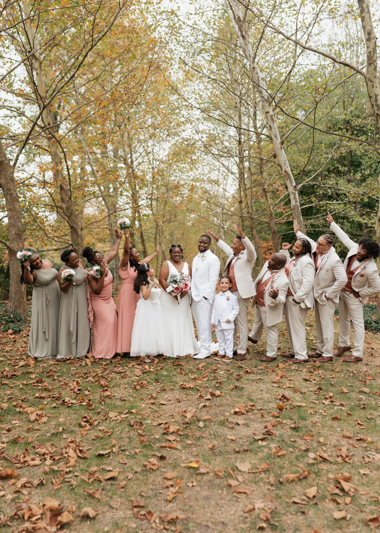 Large wedding party in white and pink attire poses on fallen autumn leaves.