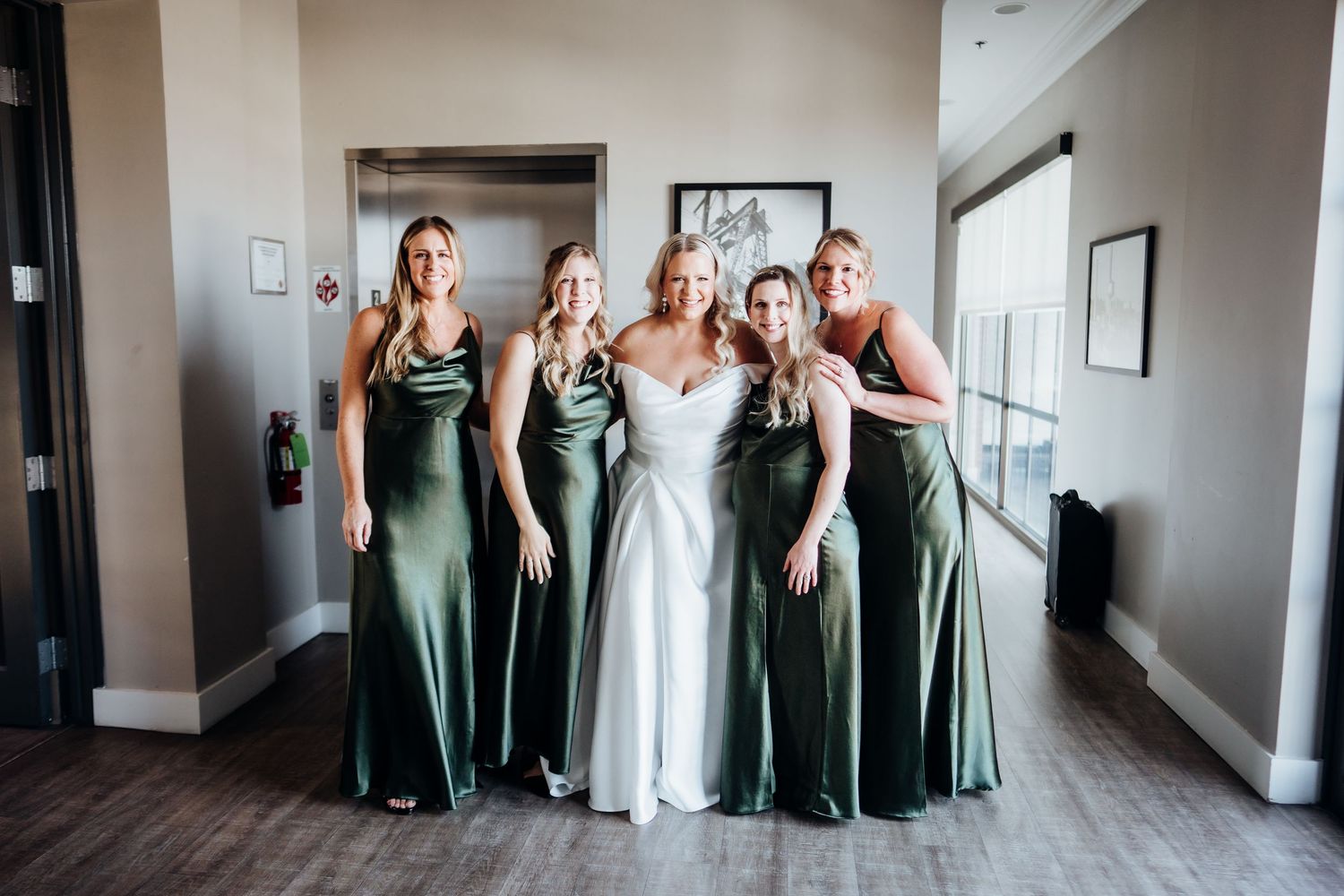 Wedding party poses in green bridesmaid dresses and white bridal gown in a modern indoor setting.