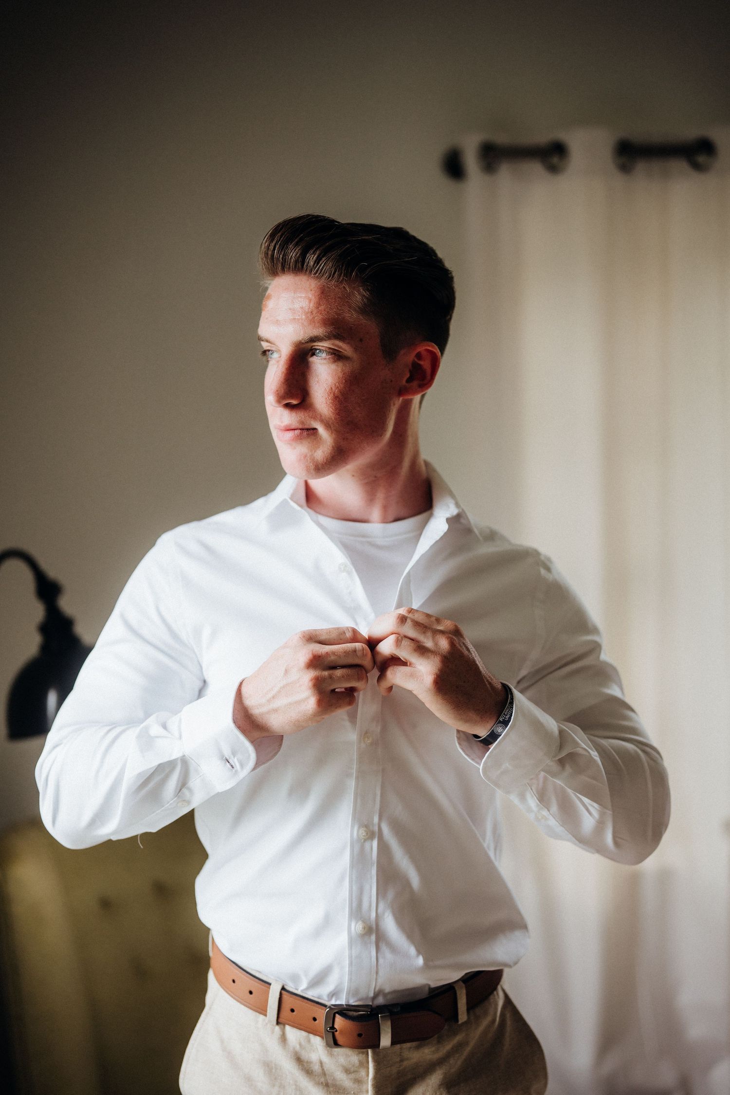 Groom getting dressed buttons up crisp white shirt in preparation for wedding day.