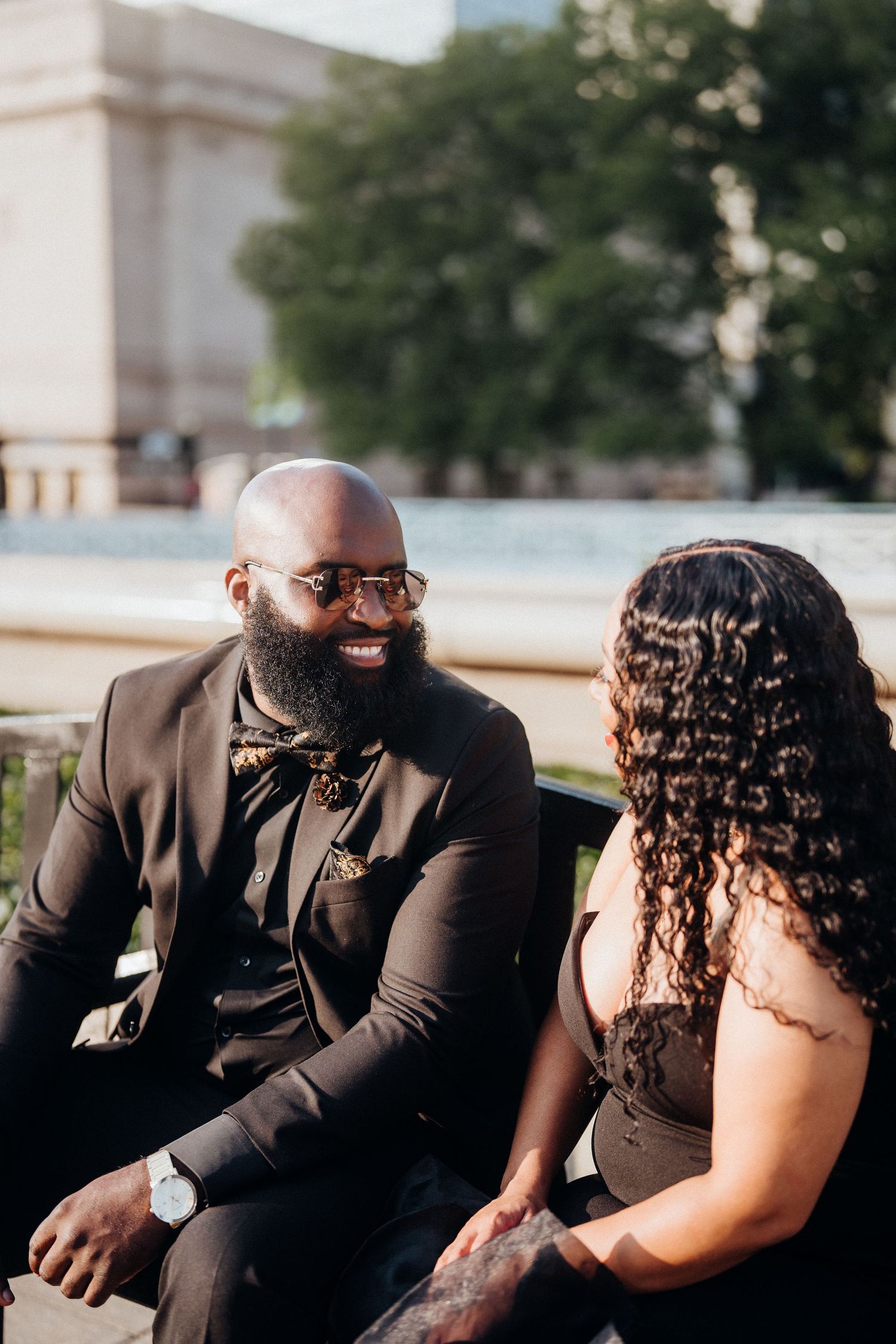 A stylish couple in formal wear sits together having a conversation outdoors.