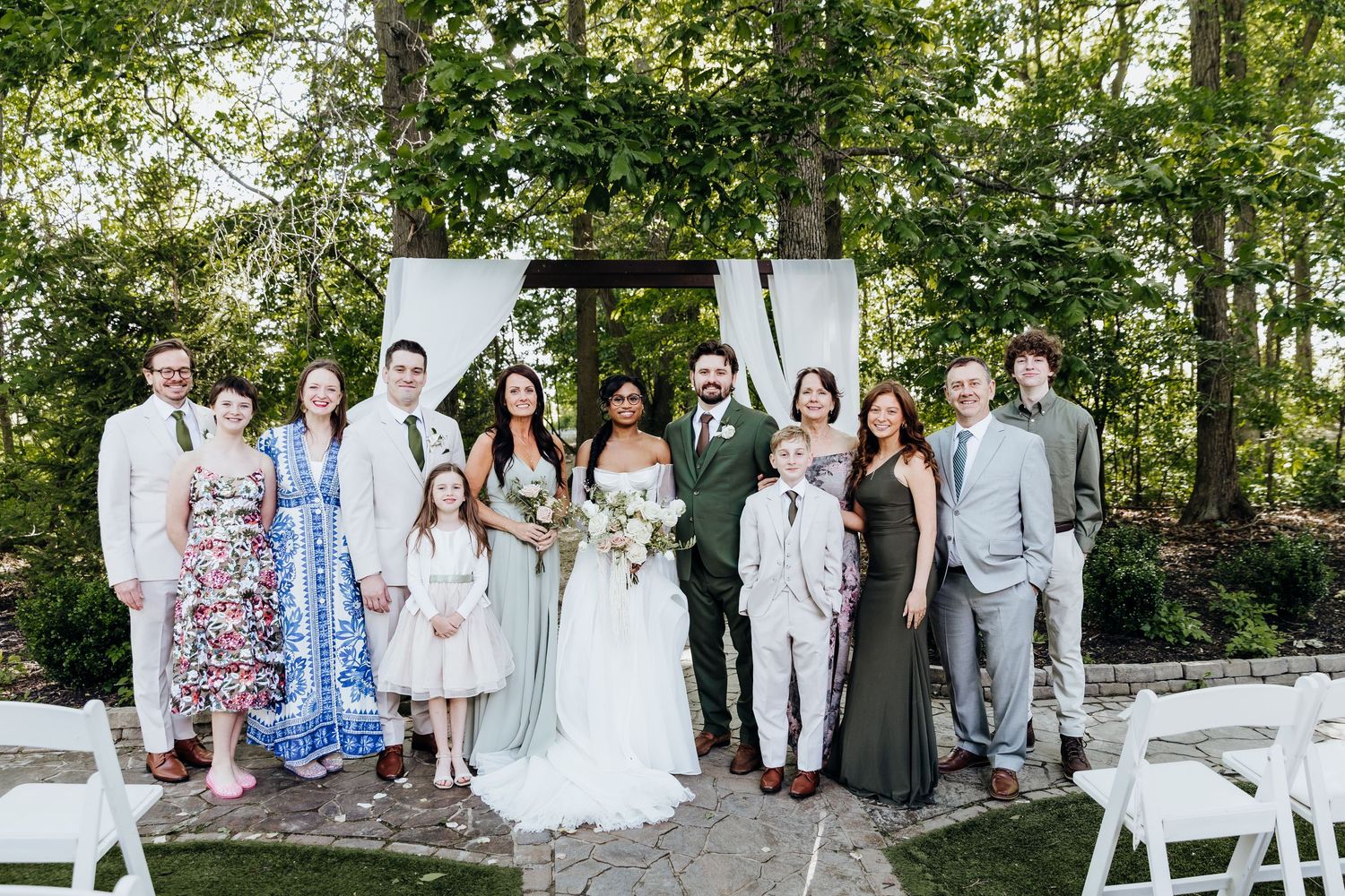 Wedding party and guests gather for outdoor group photos in garden setting.