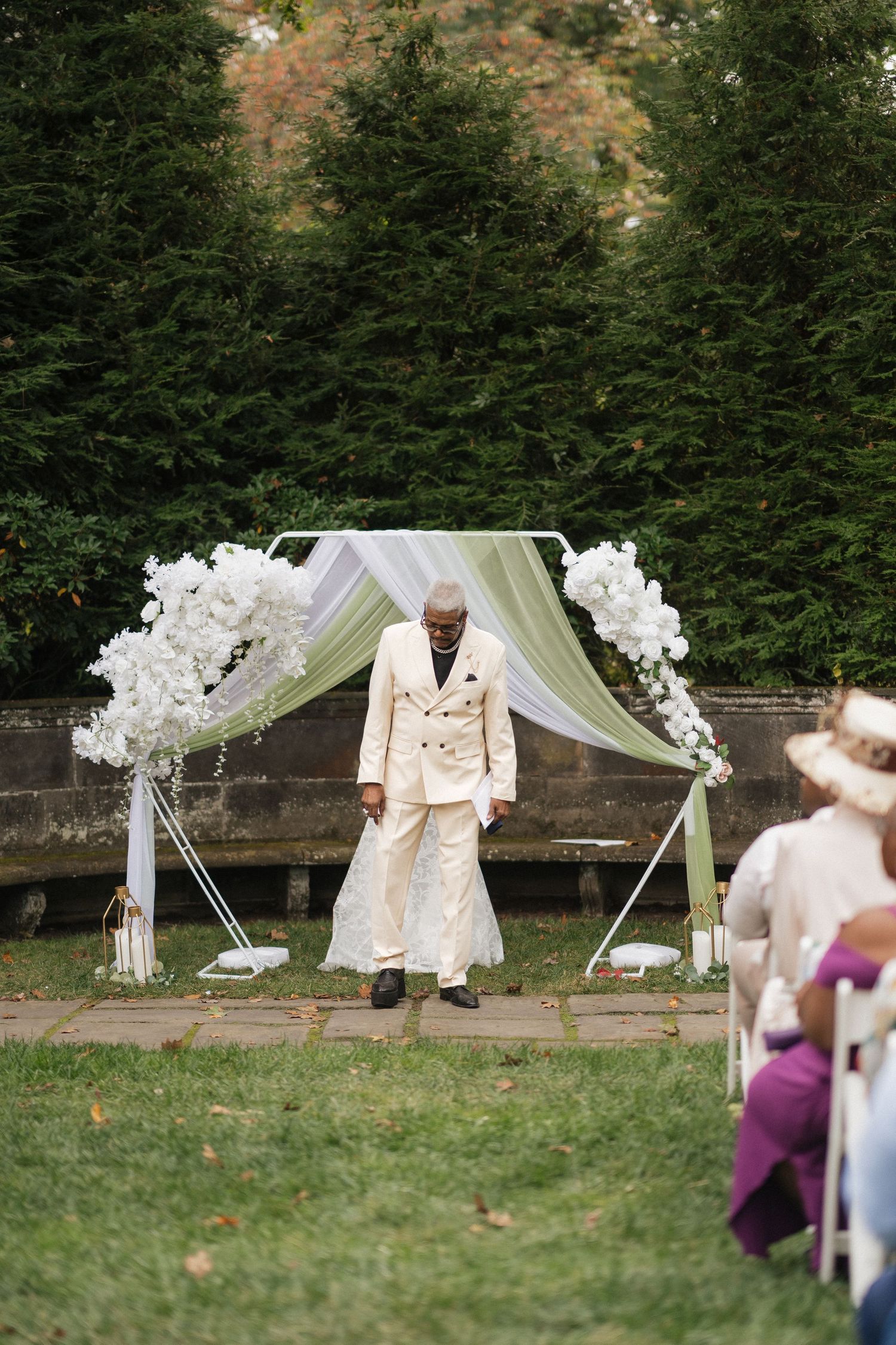 Outdoor wedding ceremony setup with white floral arch and chairs on a grassy lawn.
