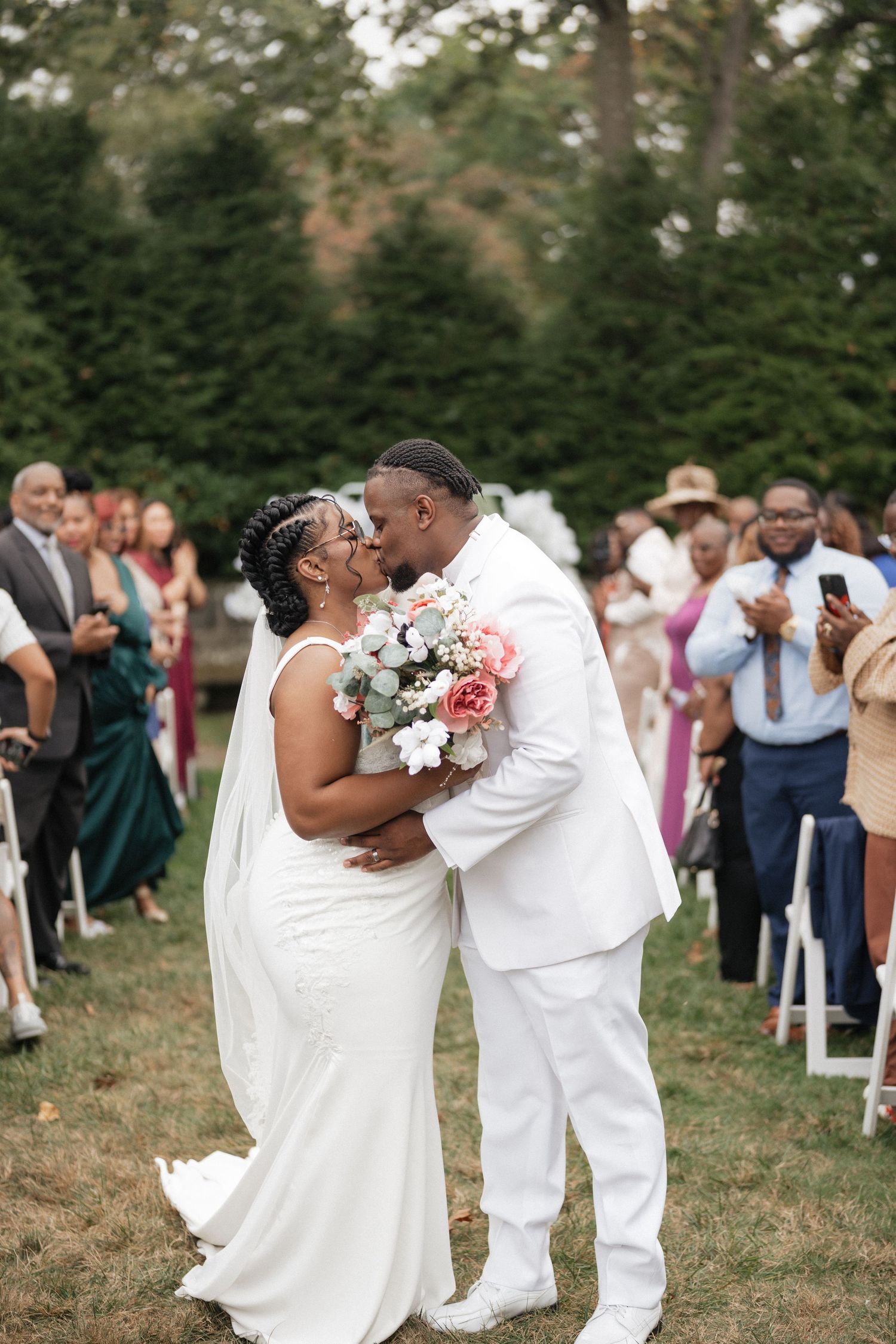 A couple shares their first kiss as newlyweds during an outdoor wedding ceremony surrounded by guests.