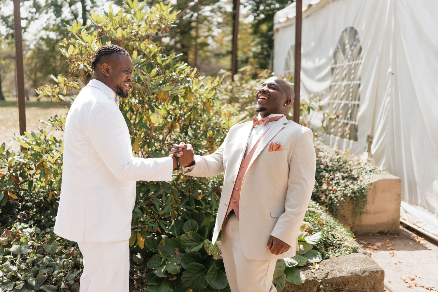 Two groomsmen in white suits share a friendly handshake outdoors.