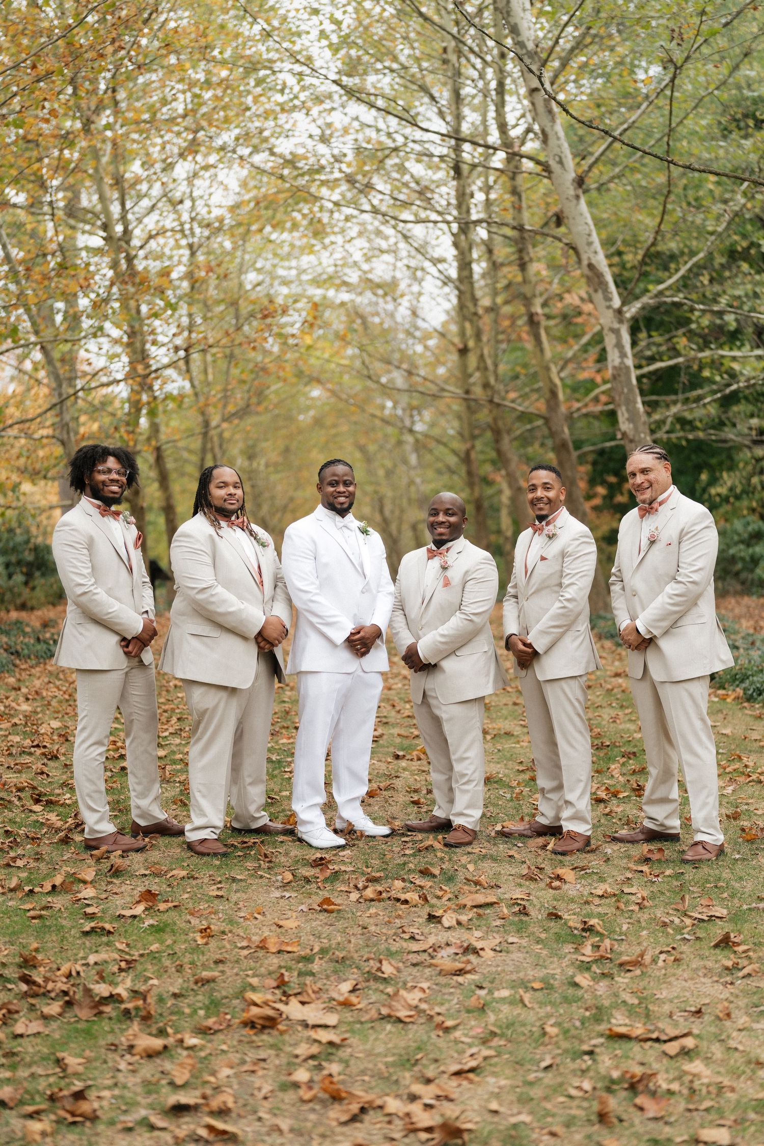Groomsmen in white suits pose casually on fallen leaves during autumn wedding.