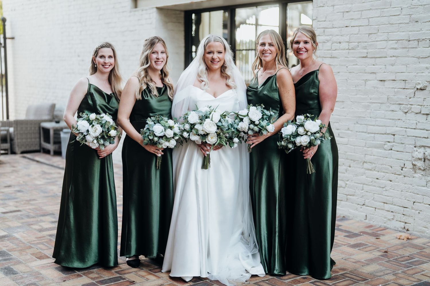 Bridesmaids in emerald green dresses pose together with white bouquets against white brick wall.