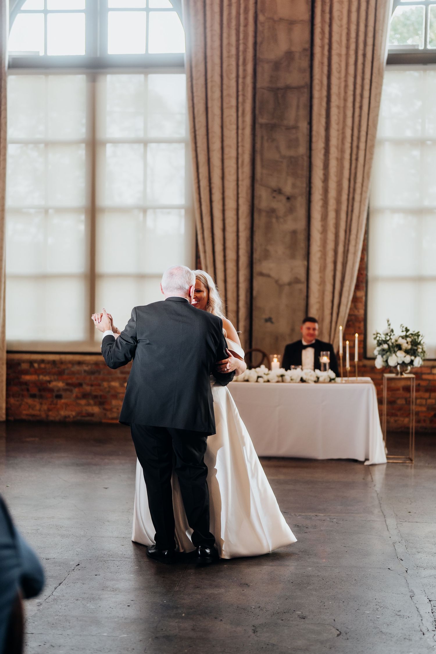 Elegant reception venue setup with white tablecloths and warm lighting through large windows.