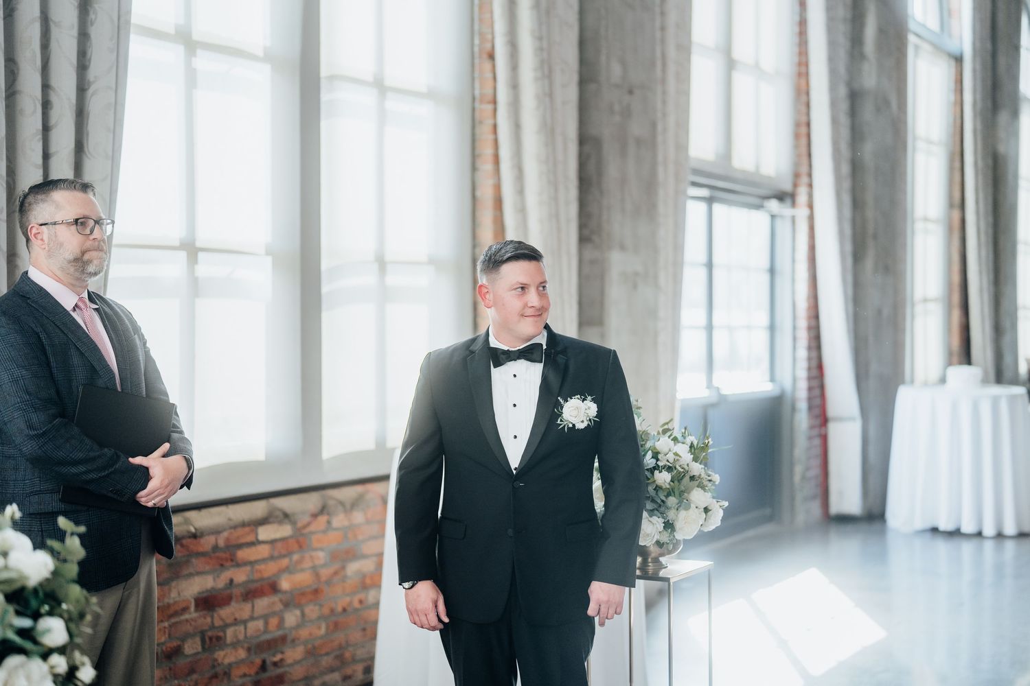 A groom in a black tuxedo stands in an elegant venue with exposed brick walls and large windows.