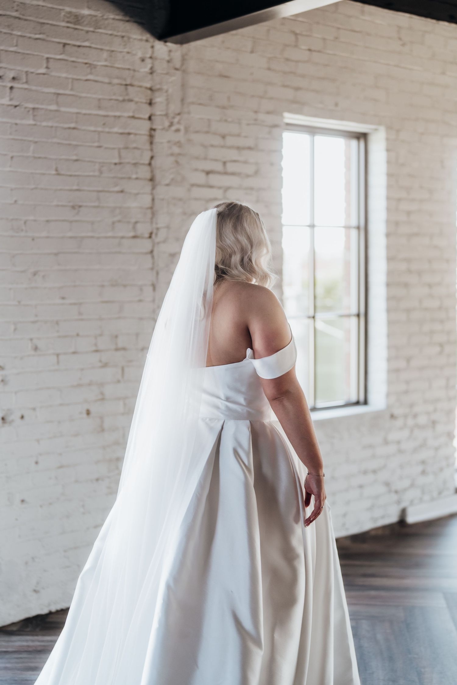 A bride in a white wedding dress poses in front of white brick walls and windows in an industrial space.