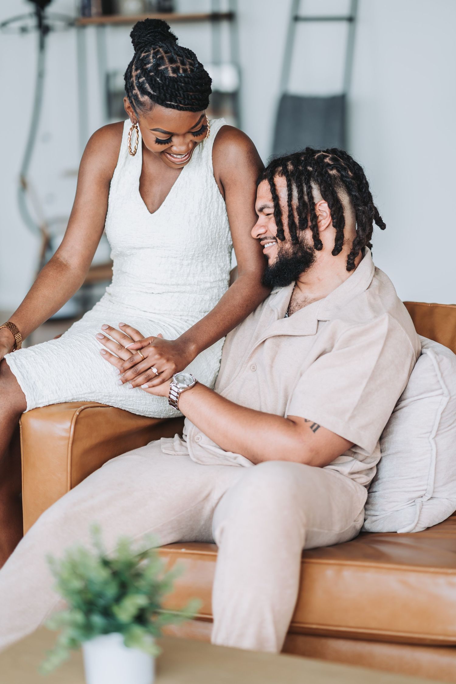A romantic couple shares an intimate moment on a tan leather couch, wearing coordinated white and beige outfits.