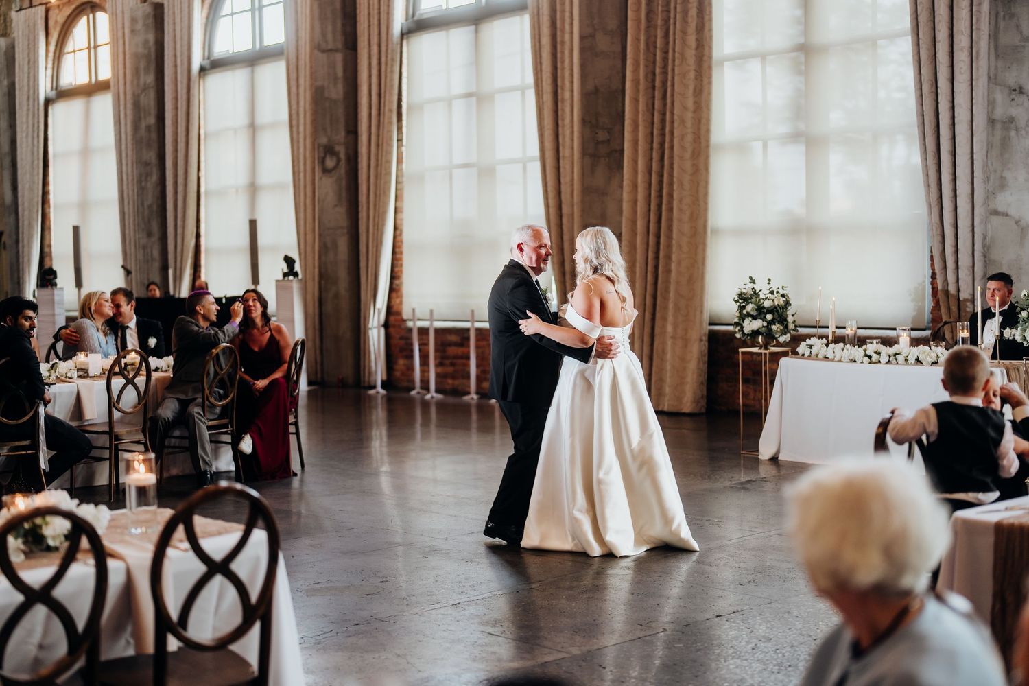Couple shares first dance in elegant ballroom with guests watching from cross-back chairs.