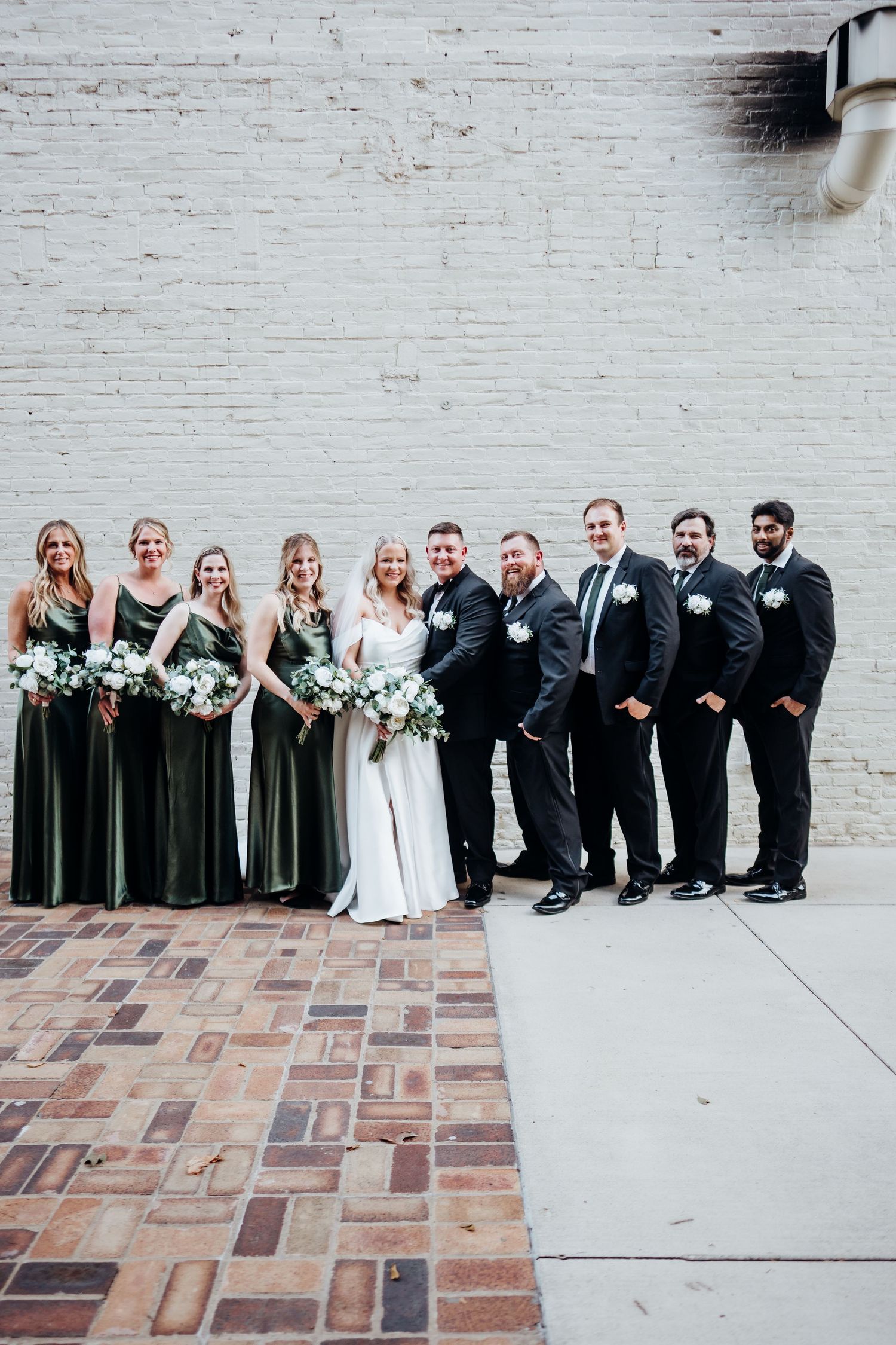 Wedding party poses against white brick wall with bridesmaids in olive dresses and groomsmen in black suits.
