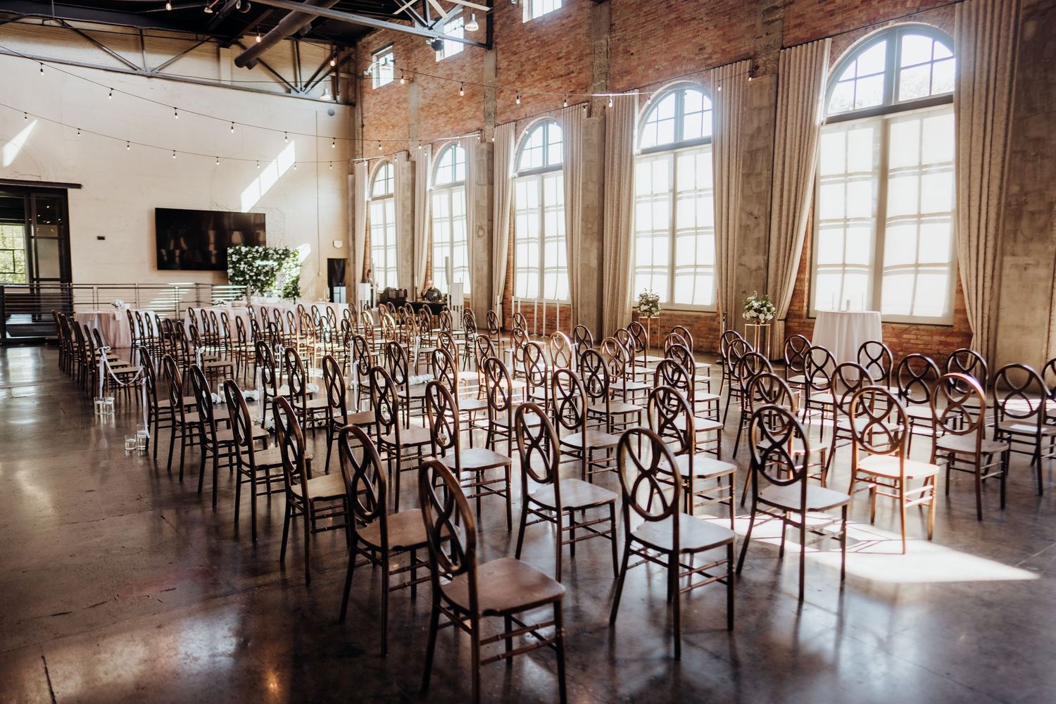 Large empty wedding venue with exposed brick walls, arched windows, and rows of wooden chairs arranged for a ceremony.
