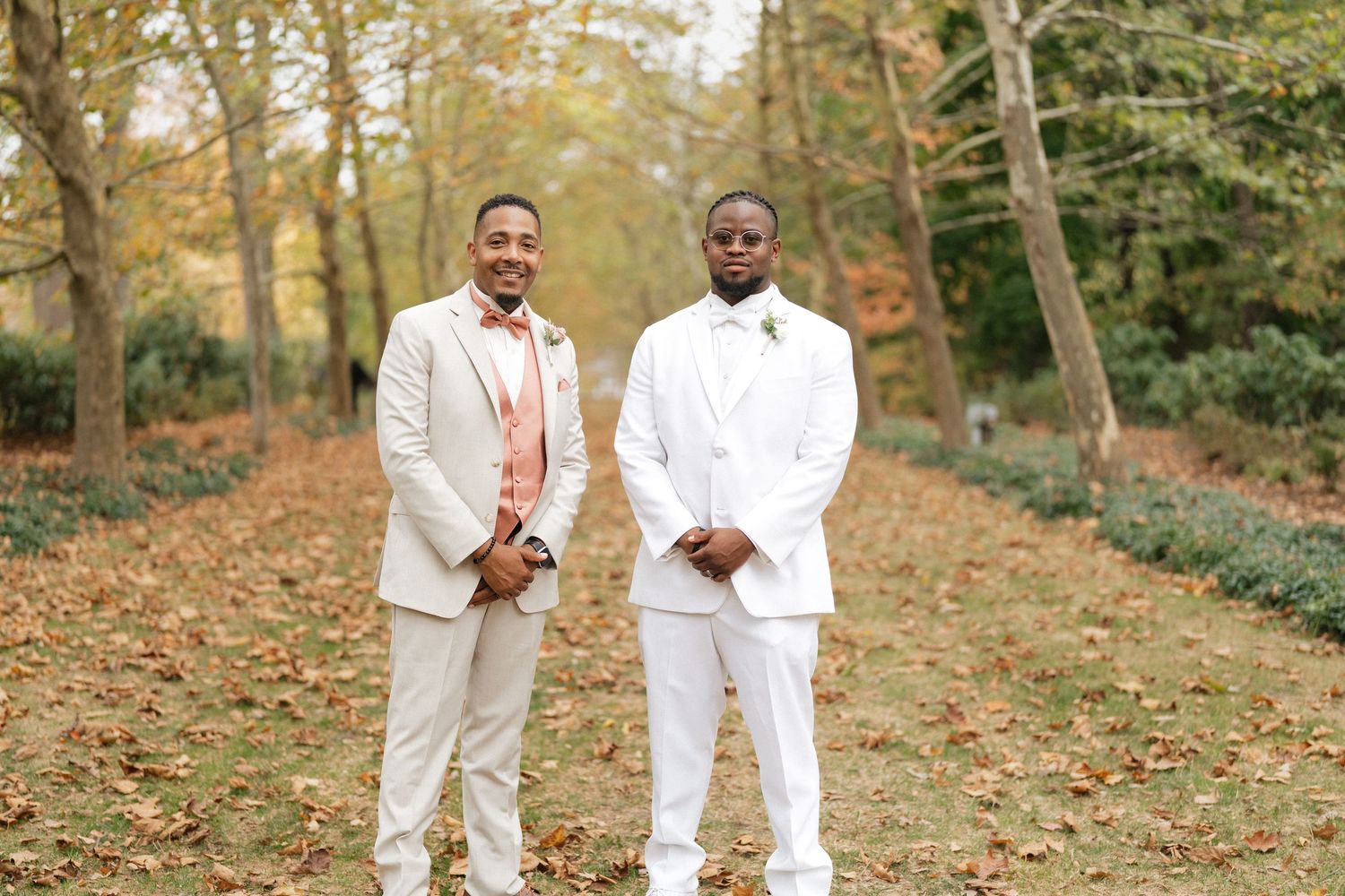 Wedding party members in white and cream suits pose on autumn grounds.