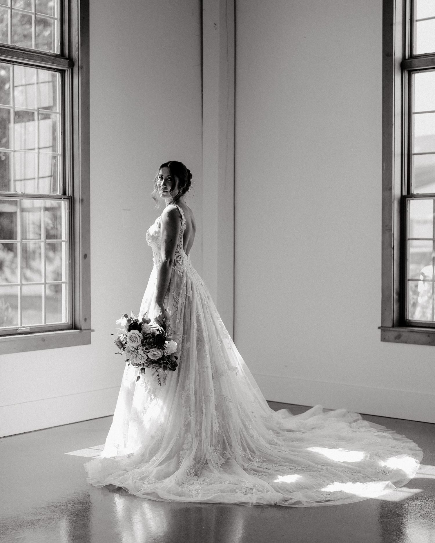 A dramatic black and white bridal portrait showing a lace gown with long train between two windows.