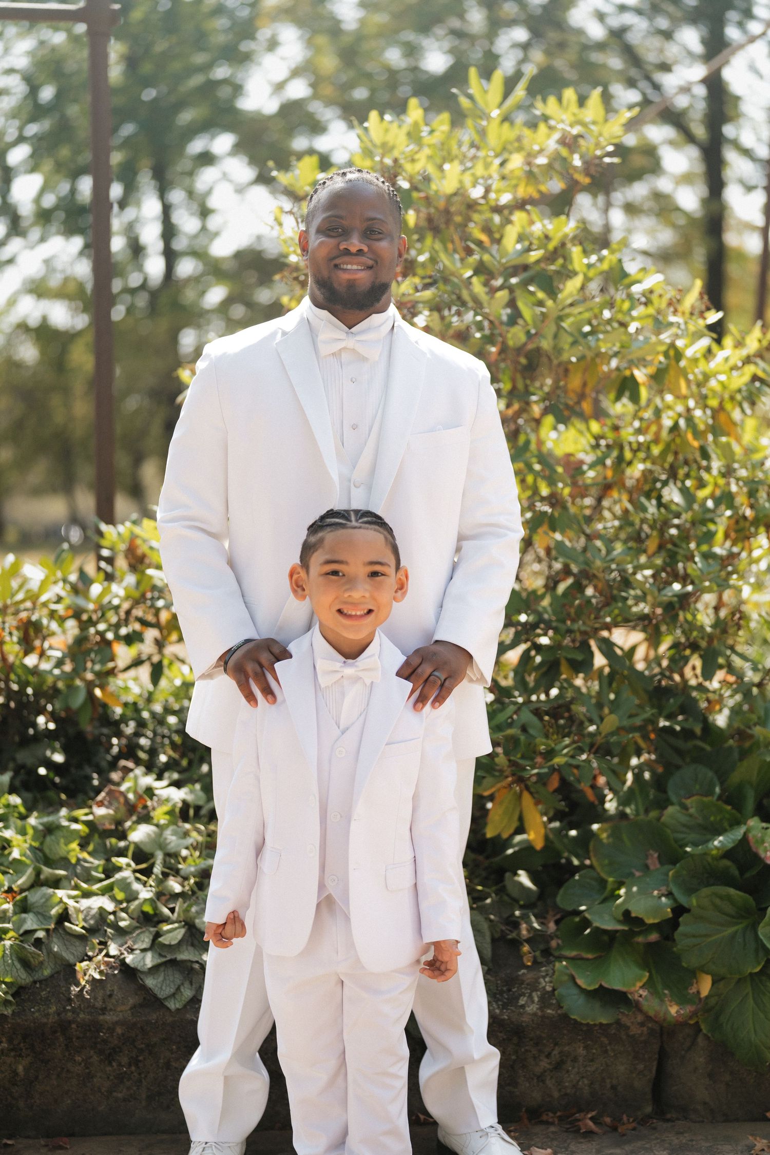 A touching father-son moment in matching white suits against a garden backdrop.