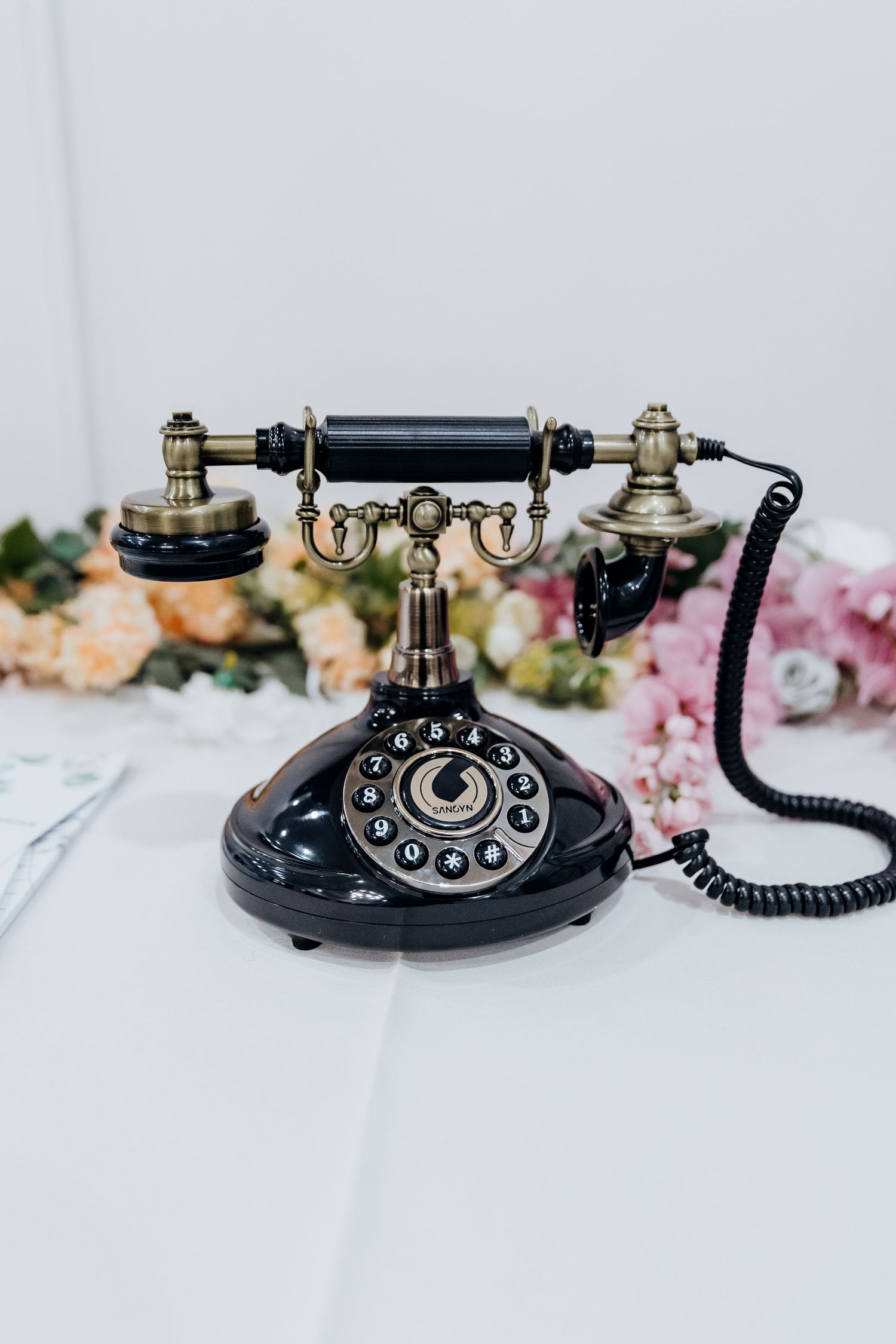 Vintage black rotary telephone with brass accents displayed with soft pink flowers on white surface.