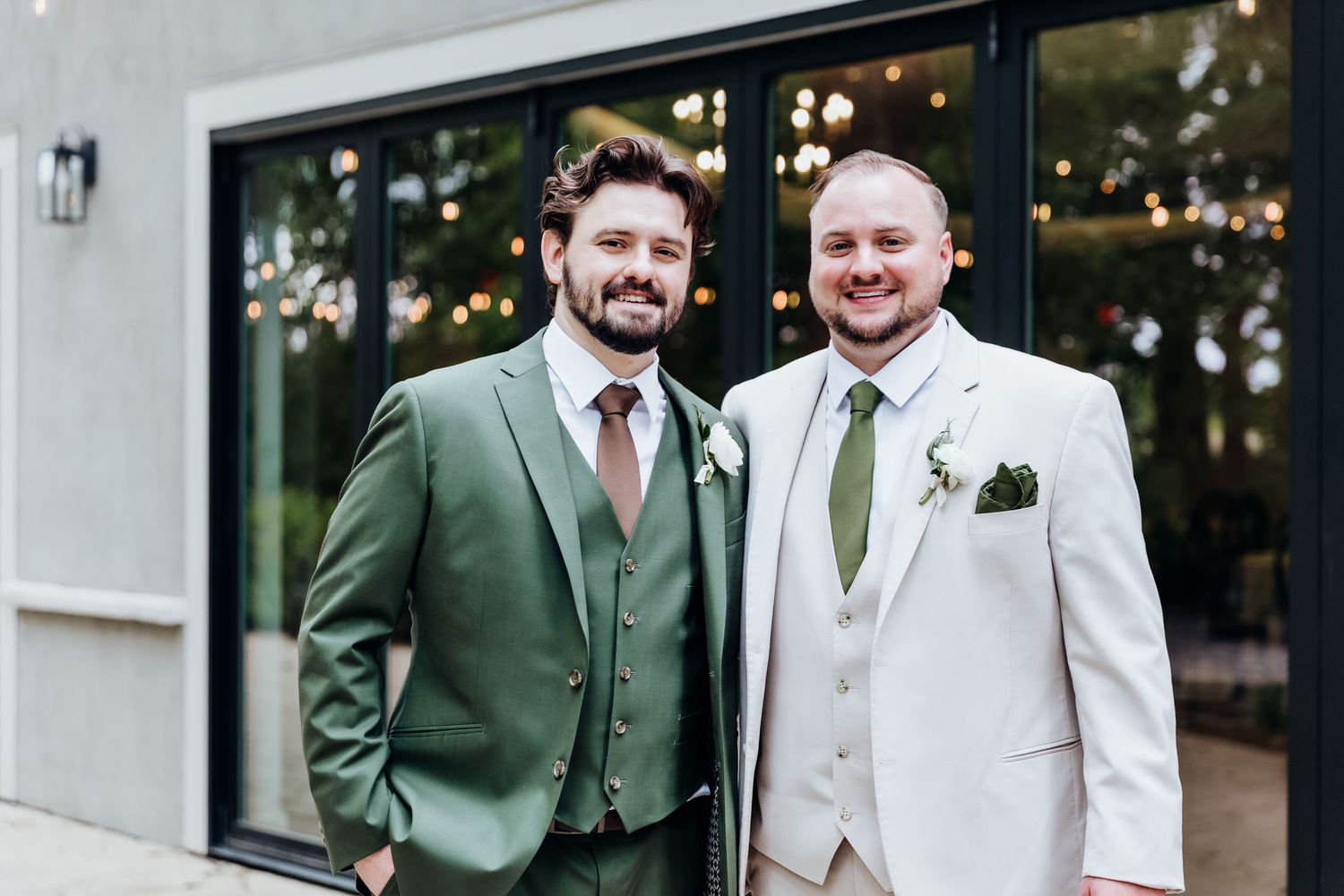 Two people wearing contrasting green and white suits smile together at an outdoor wedding venue.