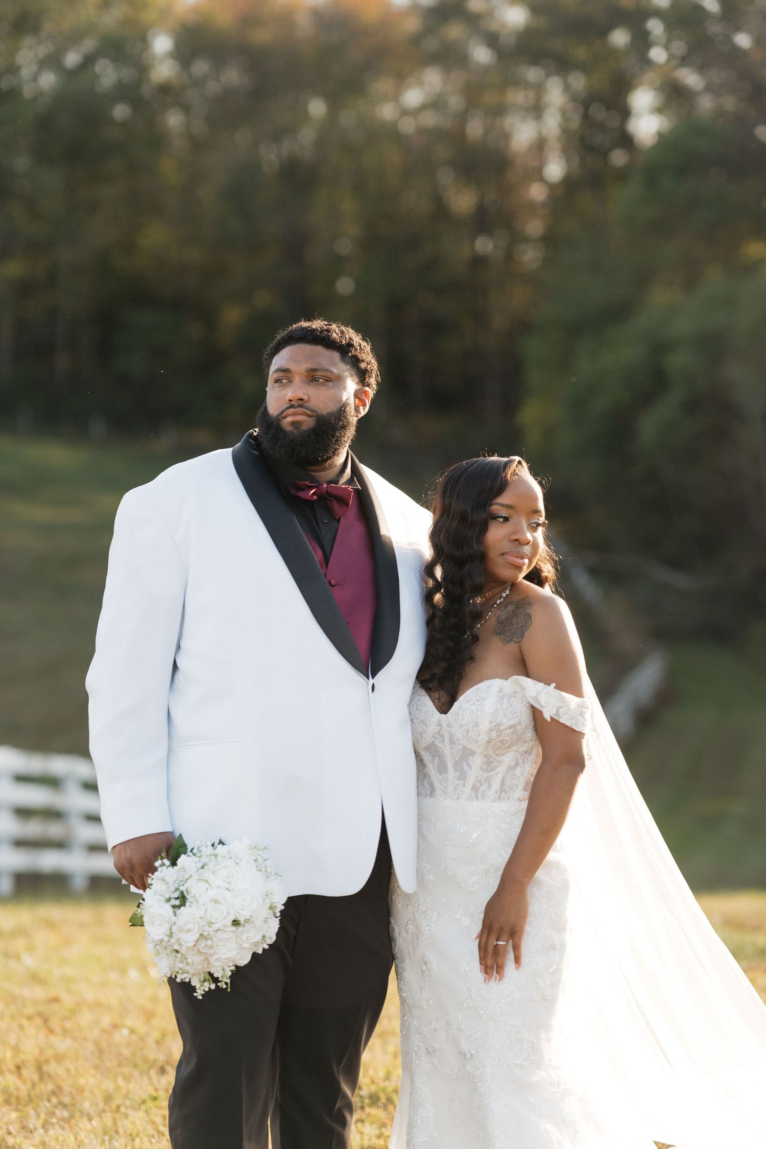 A couple in wedding attire poses outdoors, with the groom in a white jacket and the bride in an off-shoulder gown.