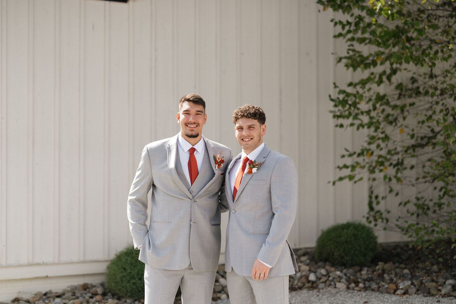 Wedding party members in light gray suits and coral ties pose together outside a building with ivy-covered walls.