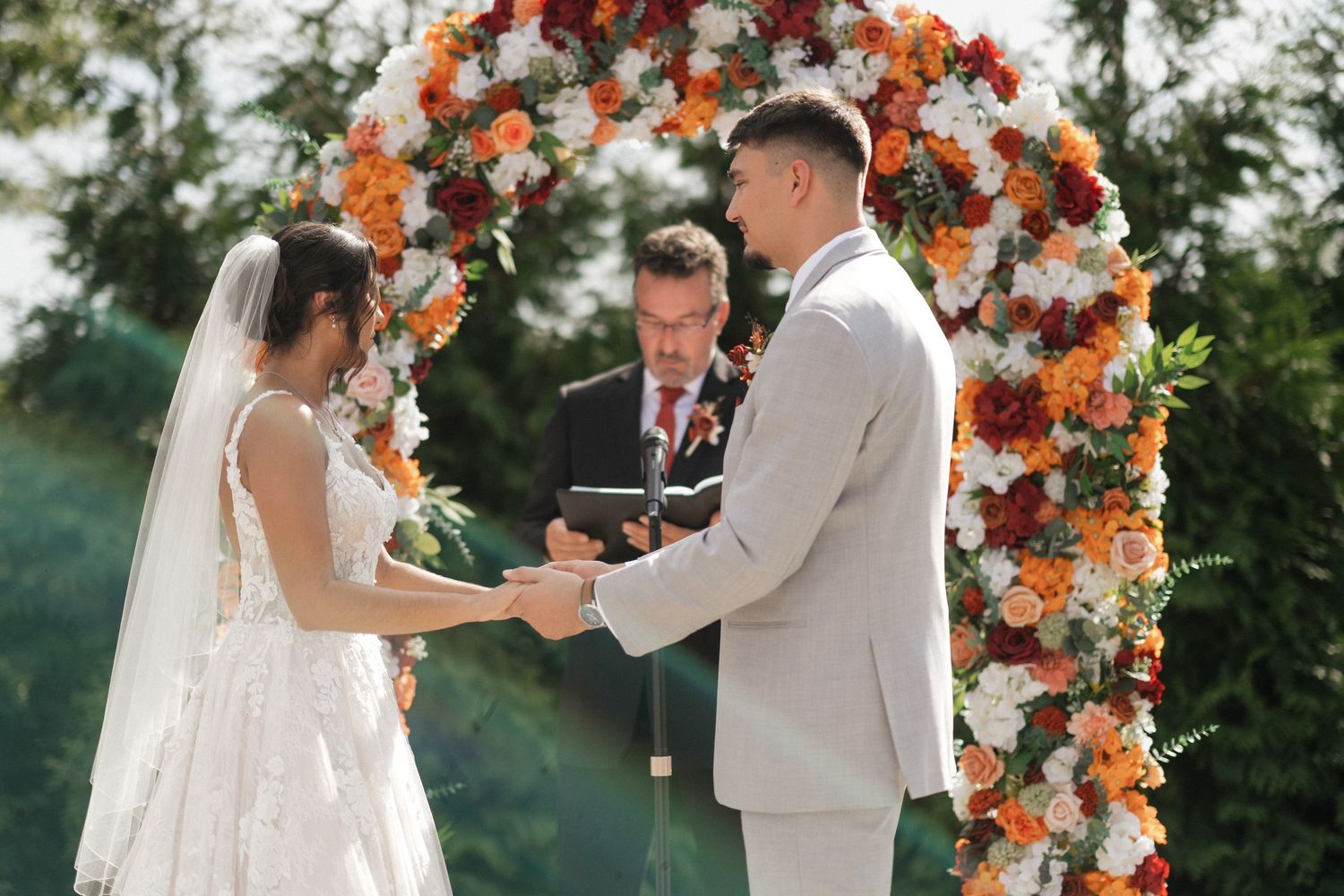 Wedding ceremony under a beautiful floral arch with orange and white flowers in an outdoor garden setting.