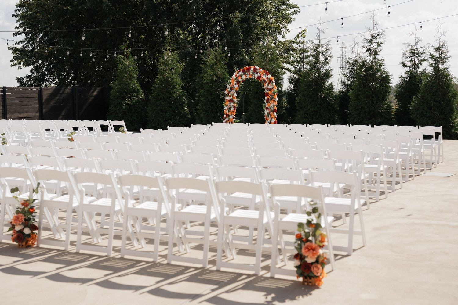 White chairs arranged outdoors for a wedding ceremony with floral arrangements and greenery in the background.