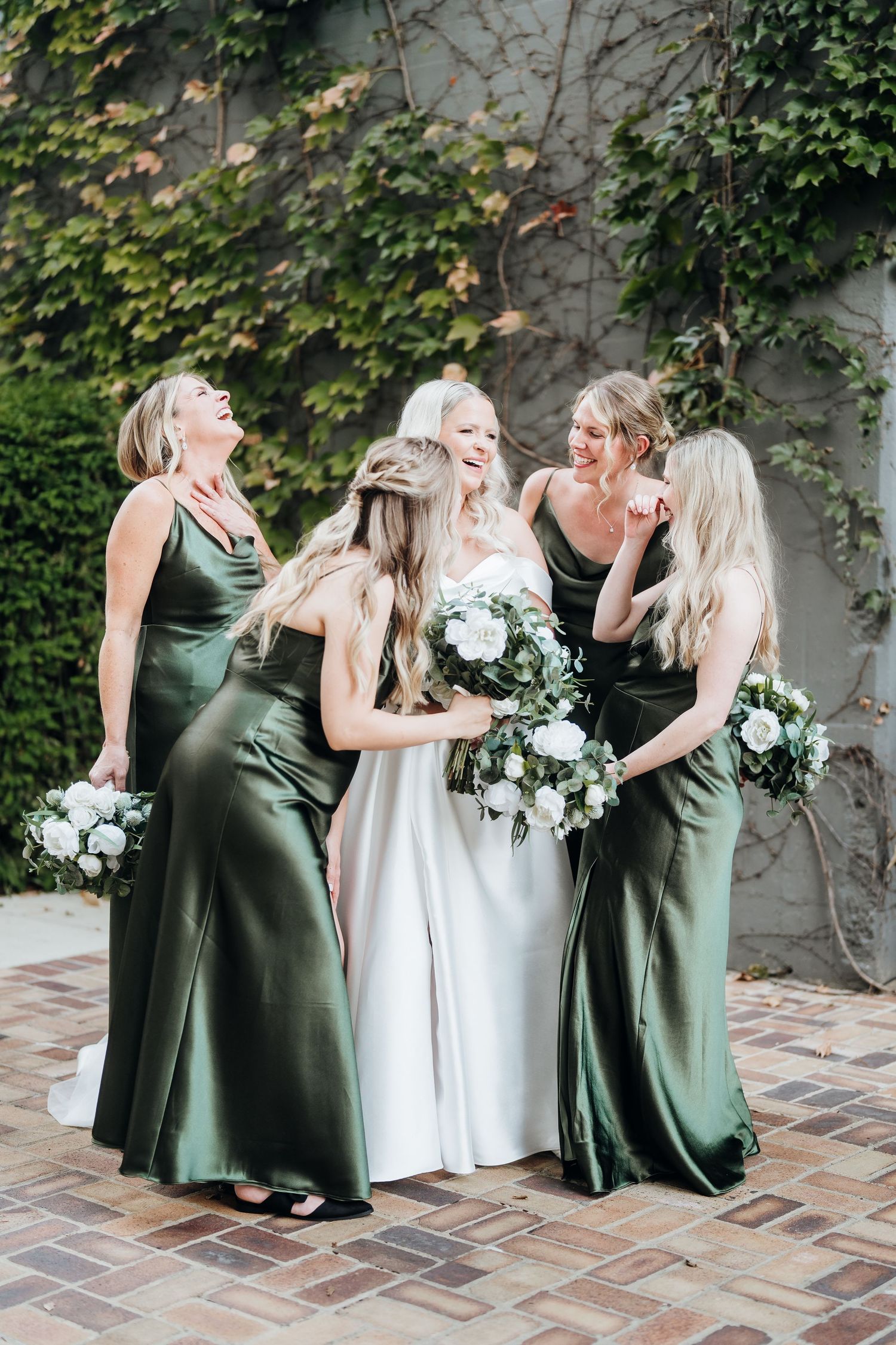 Bridesmaids in emerald green dresses gather around bride in white gown with floral bouquets.