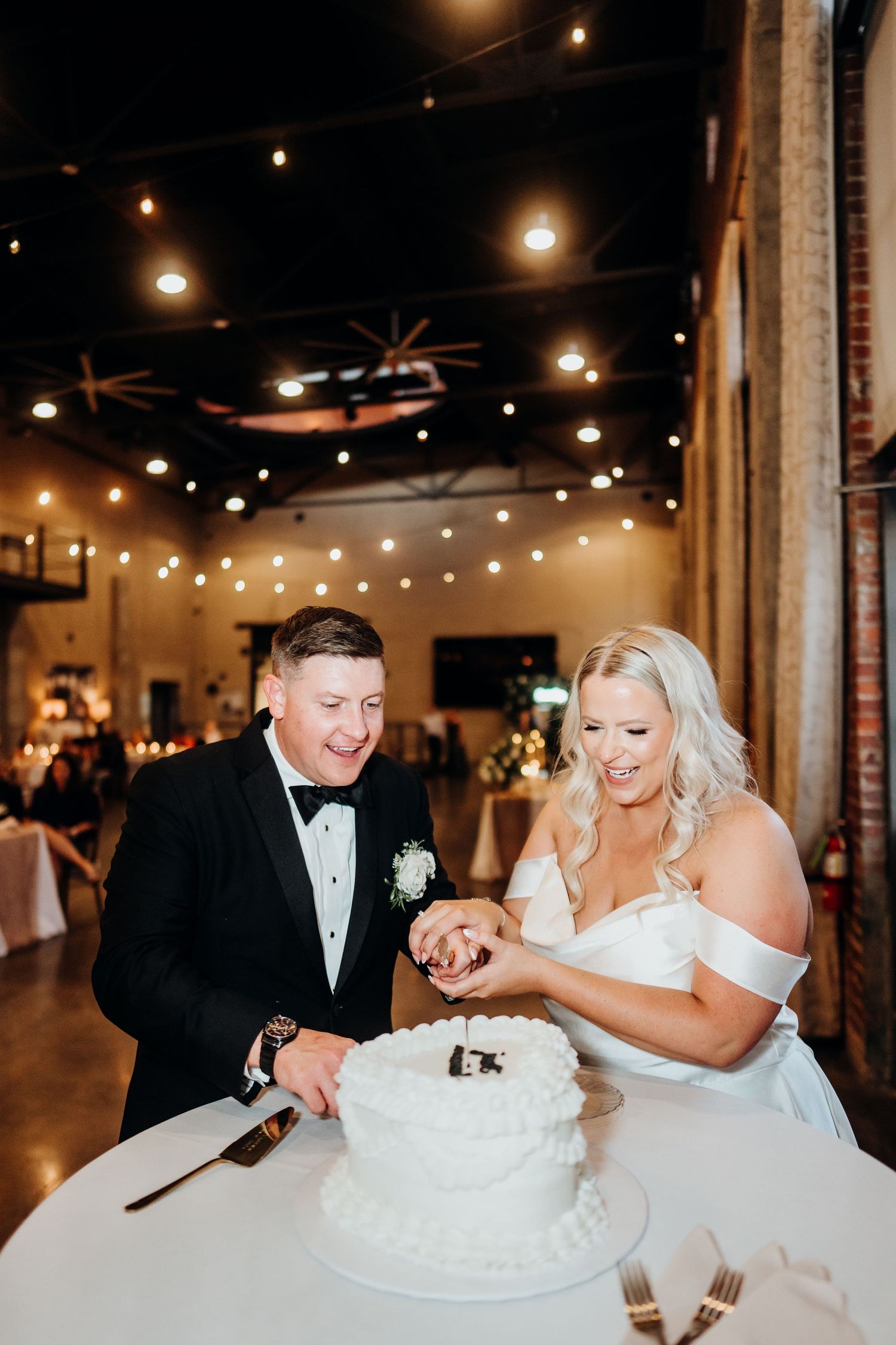 Newlyweds share a playful moment while cutting their wedding cake under warm venue lighting.