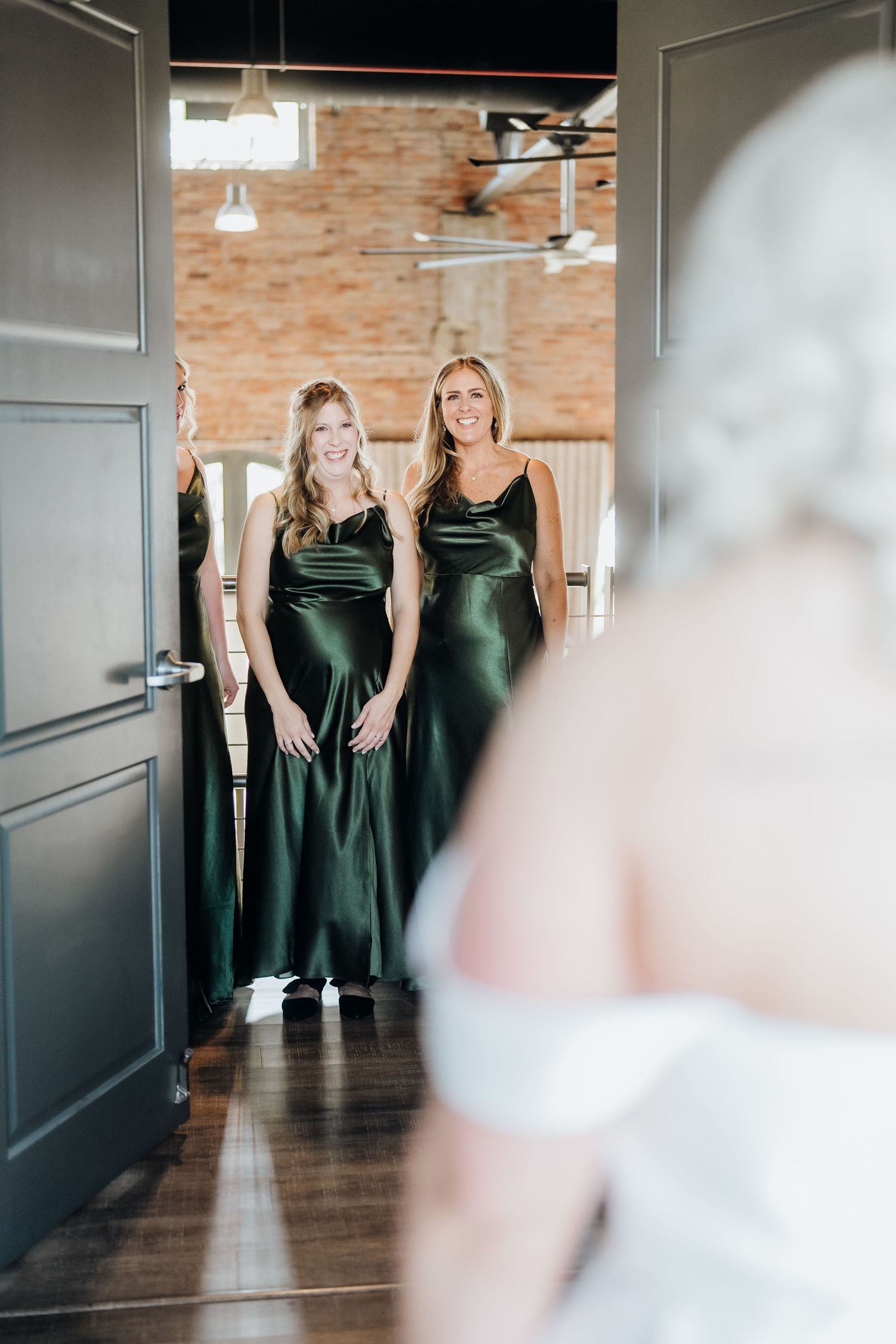 Bridesmaids in elegant dark green dresses pose in a modern venue hallway.