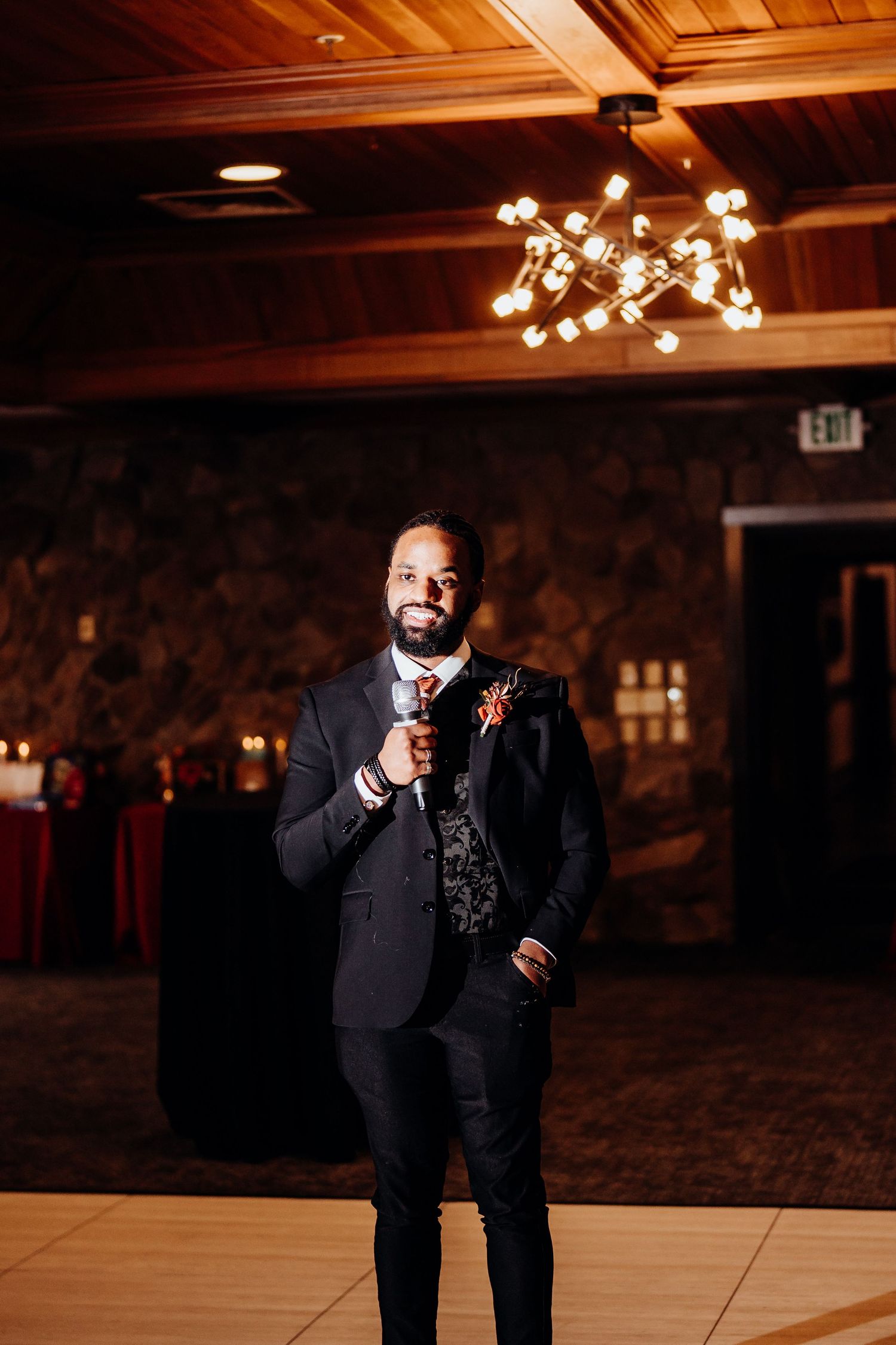 A speaker in a formal black suit addresses guests in a warmly lit reception venue with wooden ceiling.
