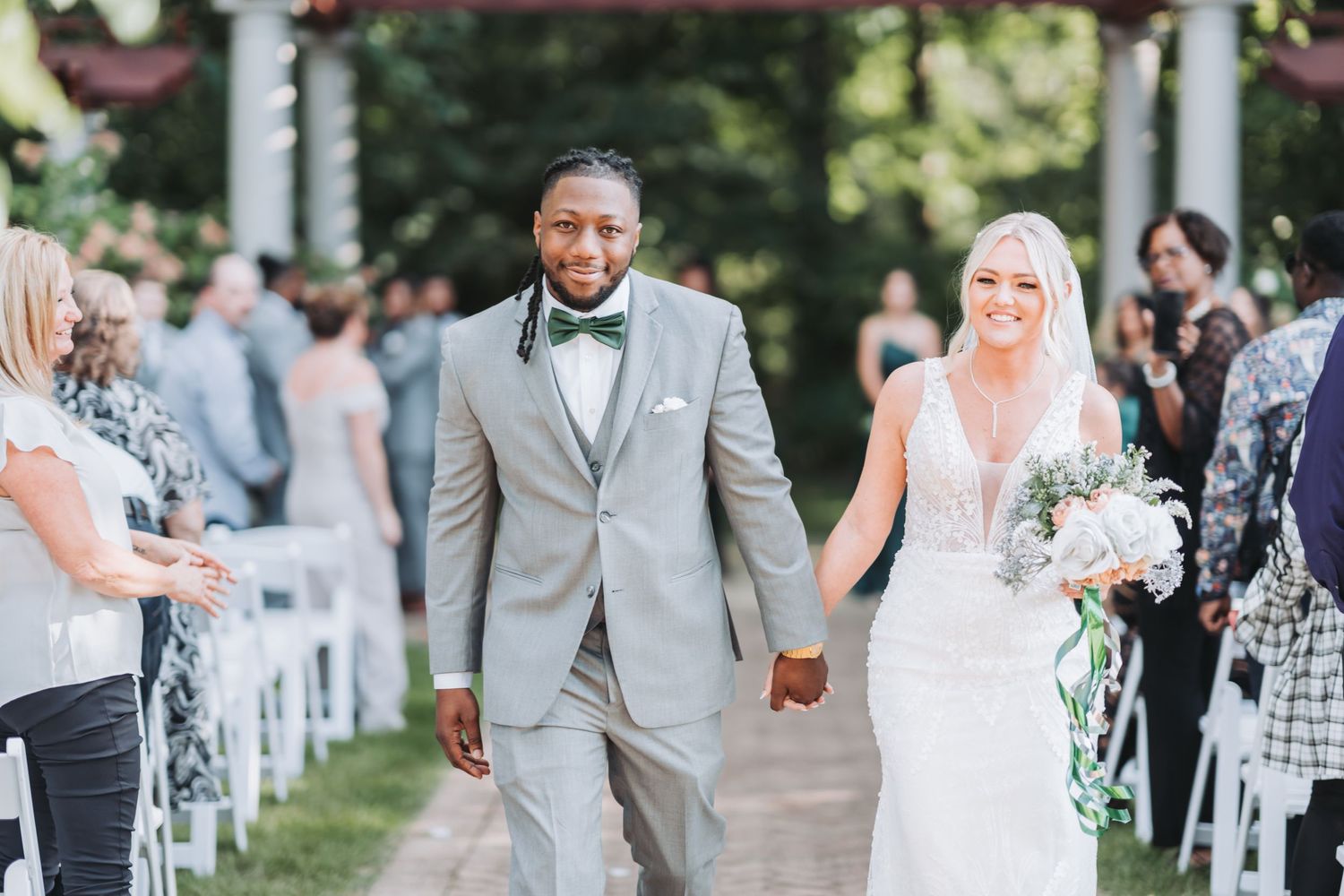 A couple walks down the aisle after their outdoor ceremony surrounded by guests and white chairs.