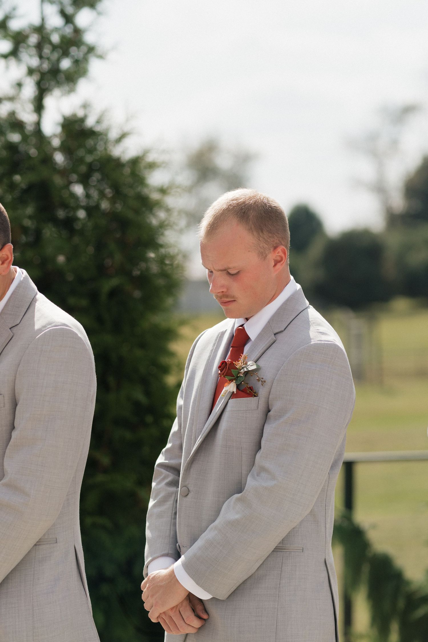 Groomsman in light gray suit with red boutonniere stands during outdoor ceremony.