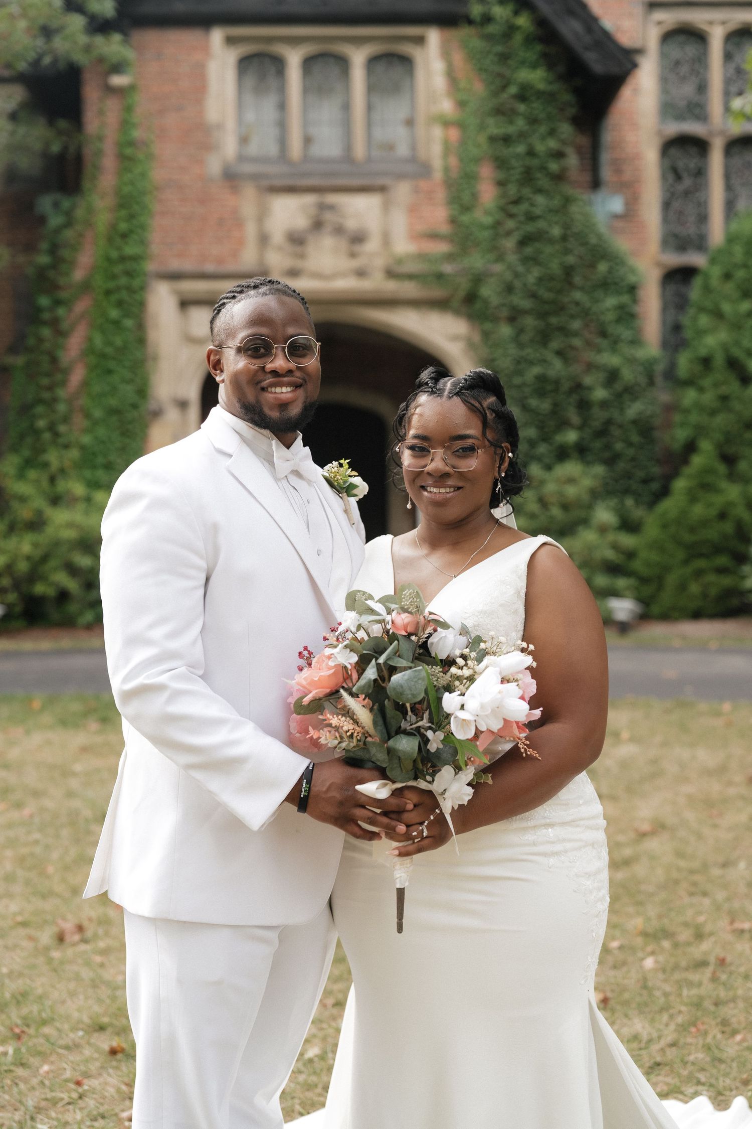 Newlywed couple in white attire sharing a joyful moment while holding a floral bouquet.