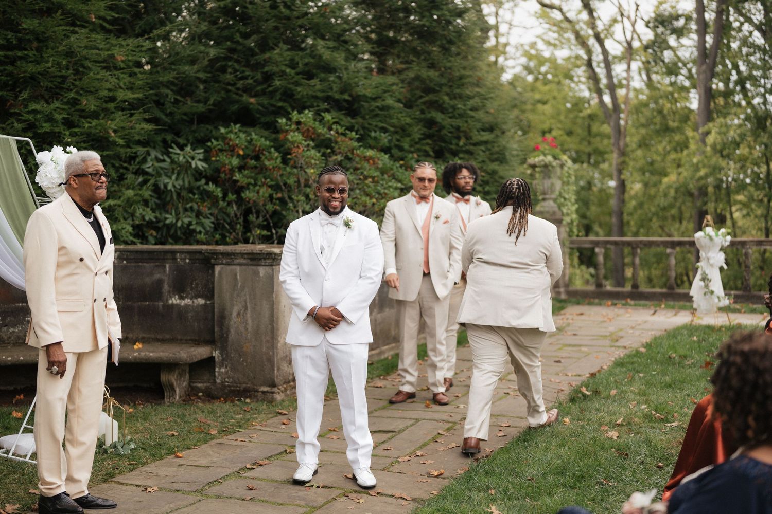 Wedding party members in white and beige attire walk along a stone pathway outdoors.