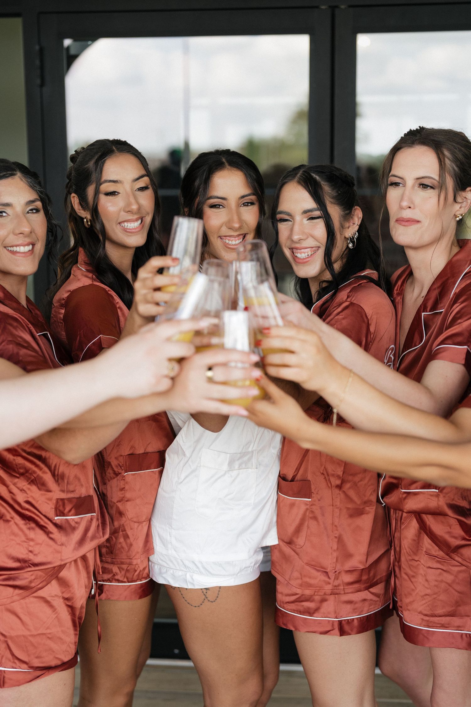 Bridesmaids in matching rust-colored silk robes raise champagne glasses together in celebration.