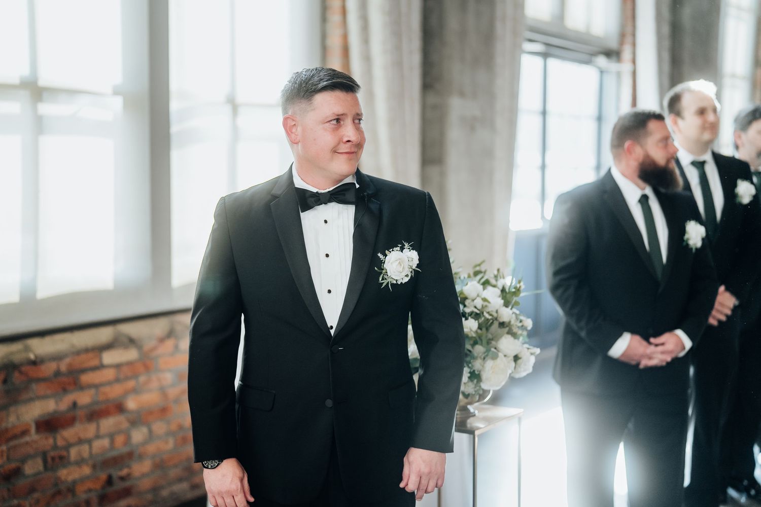 Wedding party members in formal attire gather in an industrial-style venue with large windows.