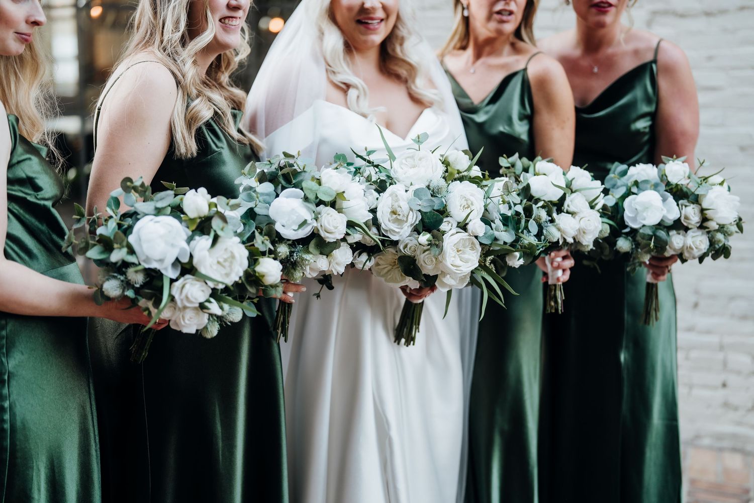 Bridesmaids in emerald green dresses holding lush white and green floral bouquets.