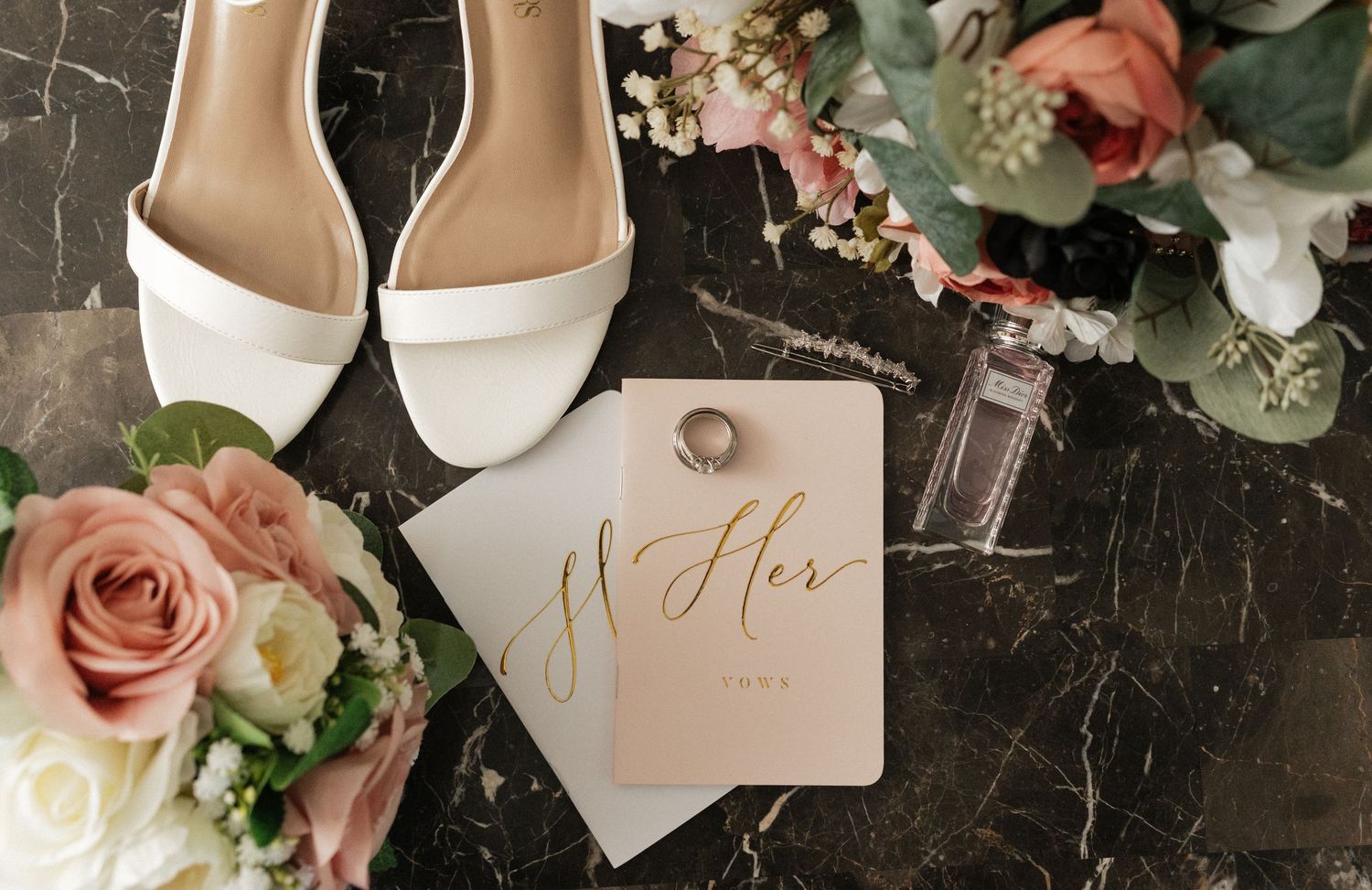 Wedding details including white shoes, pink roses, and elegant stationery arranged on dark marble surface.