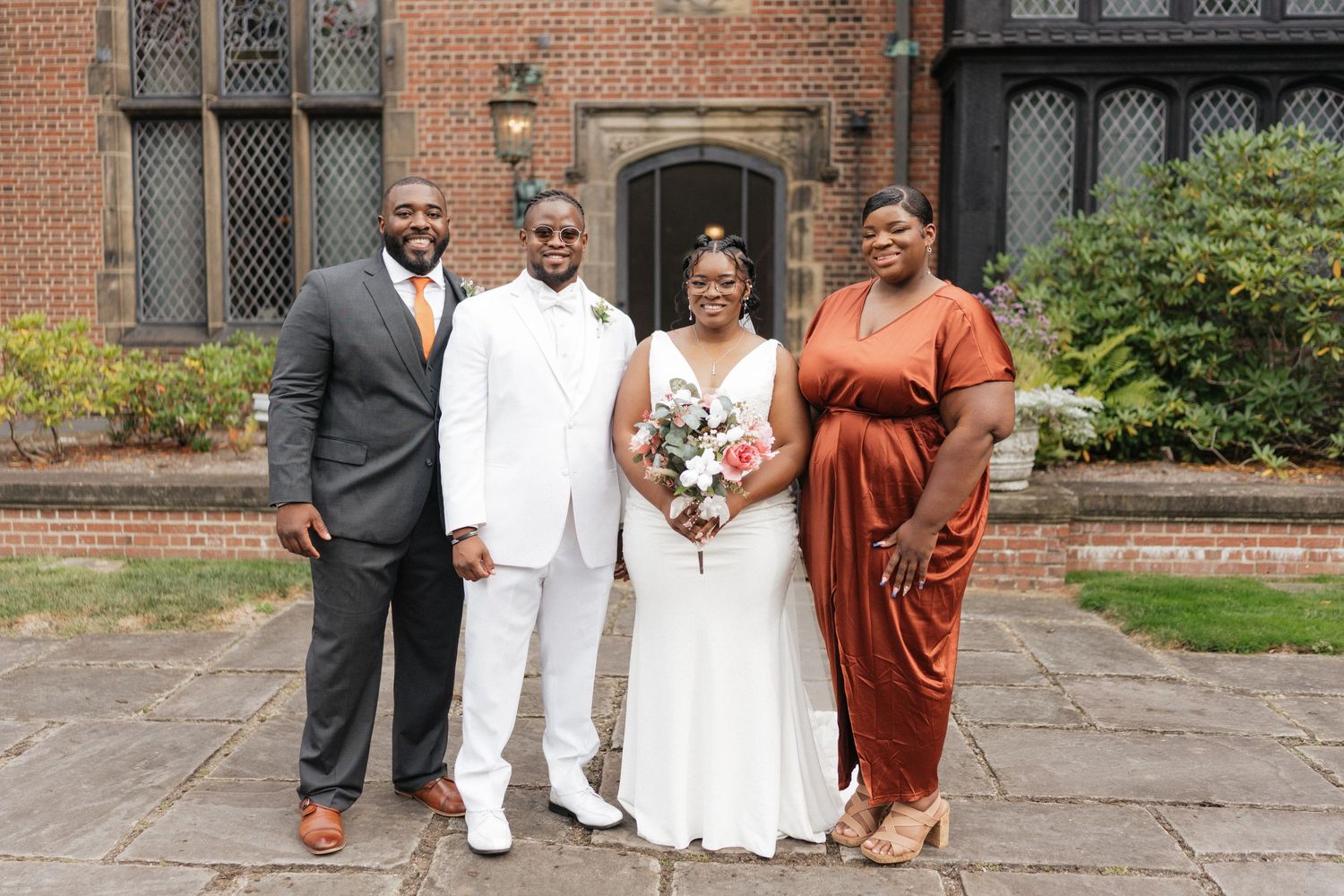 Wedding party members in white and burgundy attire gather for formal photos outside a brick church.