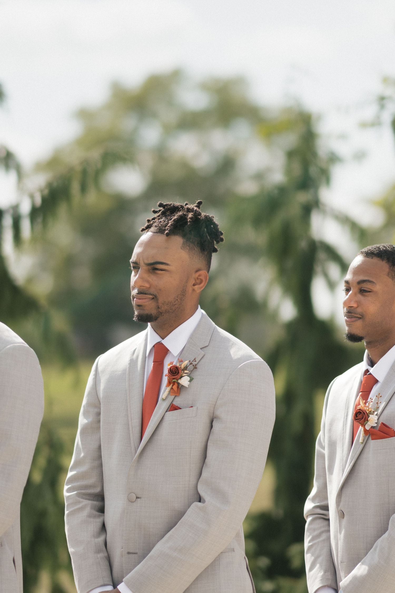 Groomsmen in light gray suits with coral ties stand in line during outdoor ceremony.