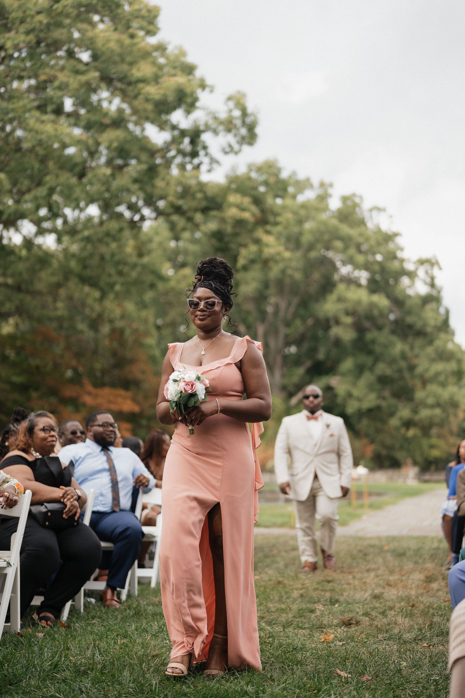 Wedding ceremony guests seated outdoors on a grassy hill with trees in the background.