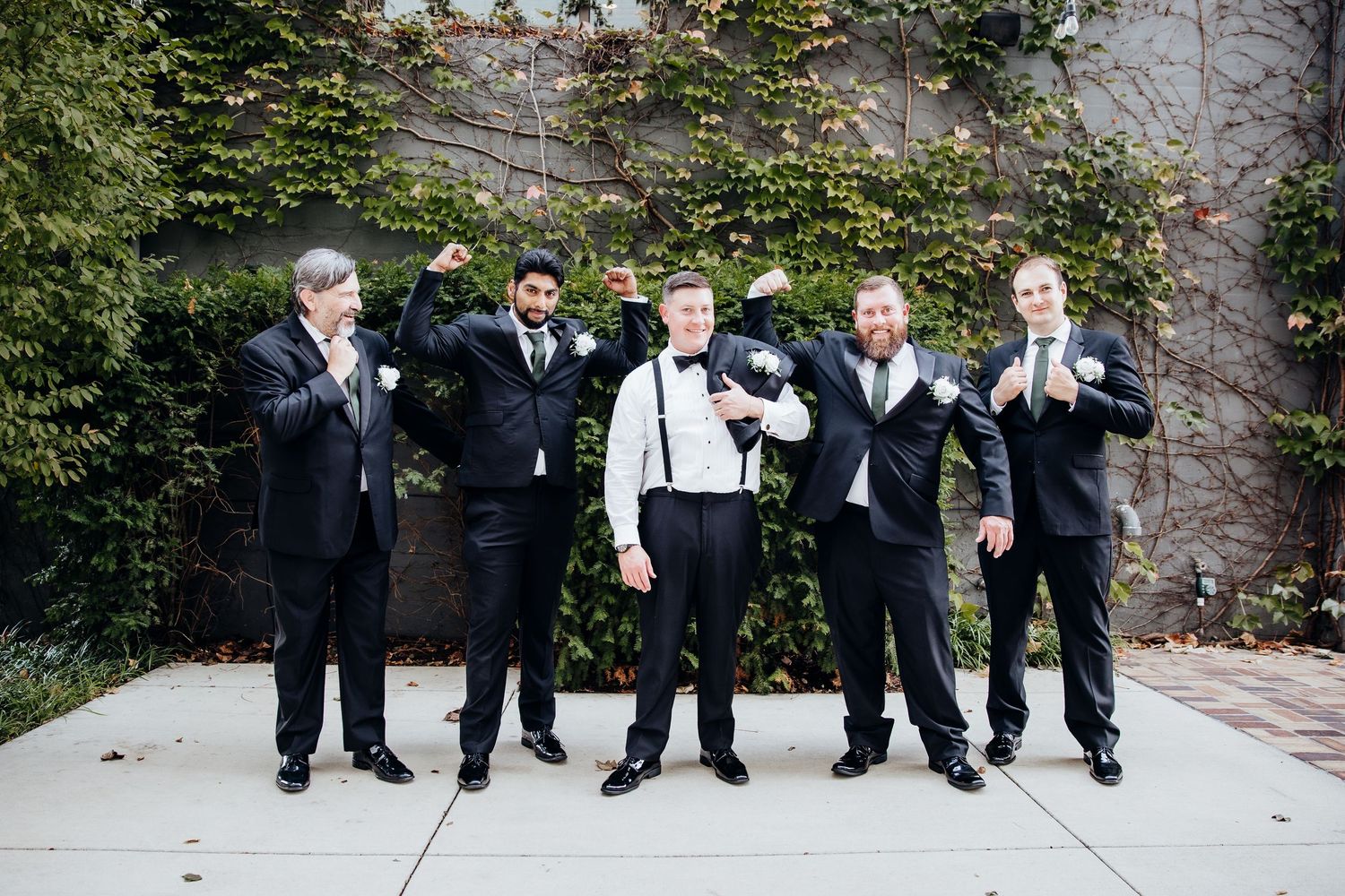 Groomsmen in black suits sitting casually outdoors against stone wall.