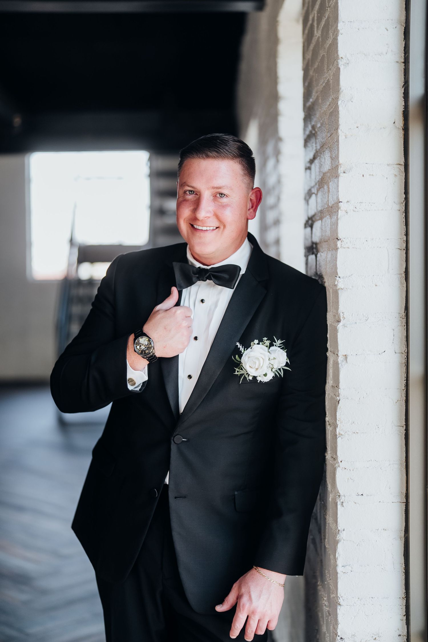 Groom in classic black tuxedo poses against industrial white brick wall.