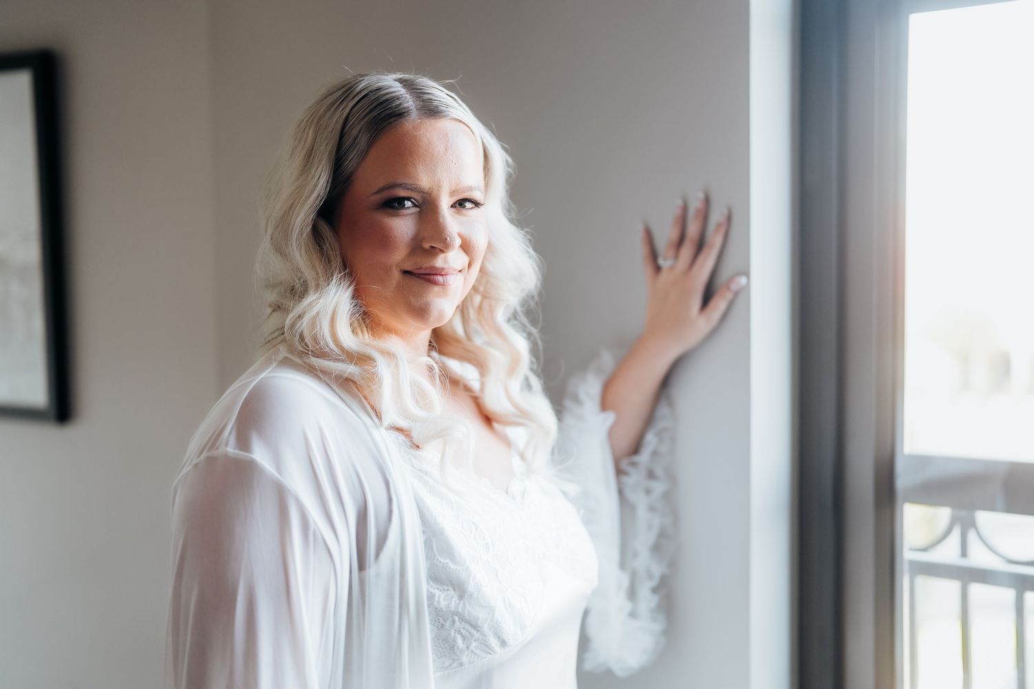 Bride in a flowing white wedding dress poses elegantly by a bright window in a modern interior setting.