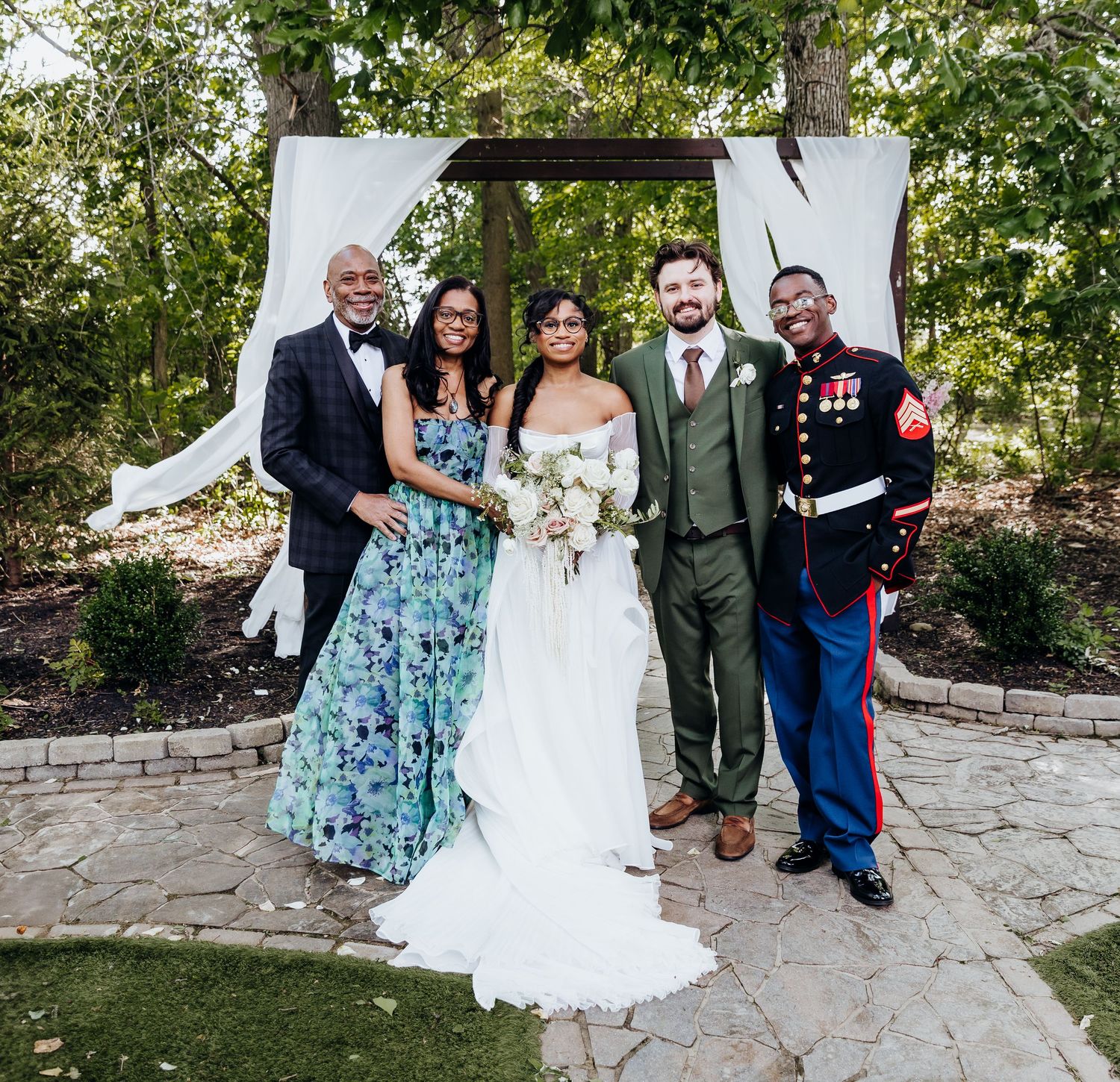 Wedding party poses under white draped arch with military service member and guests in formal attire.