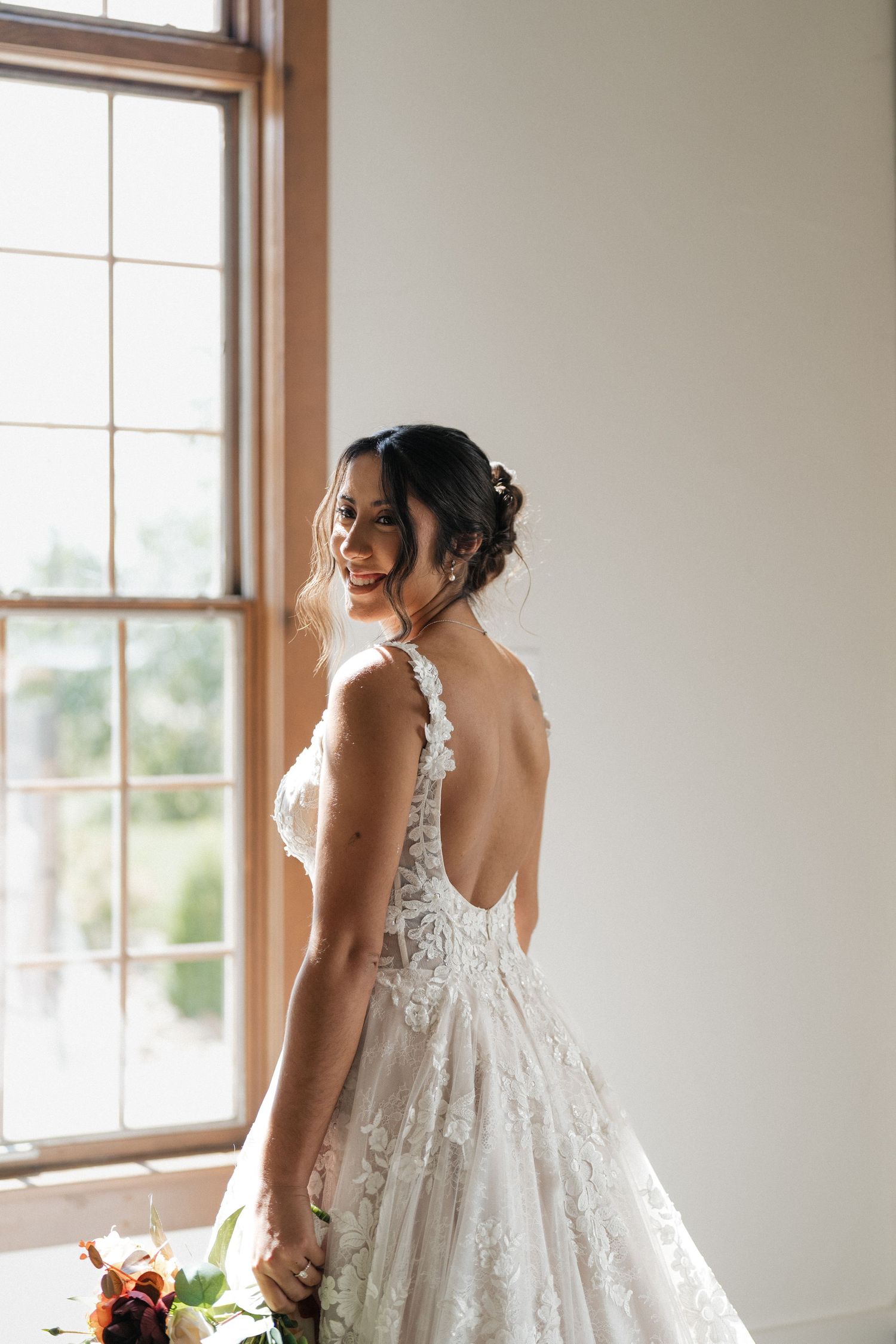 A bride in a delicate lace wedding gown stands by large windows in a sunlit room, creating an elegant silhouette.